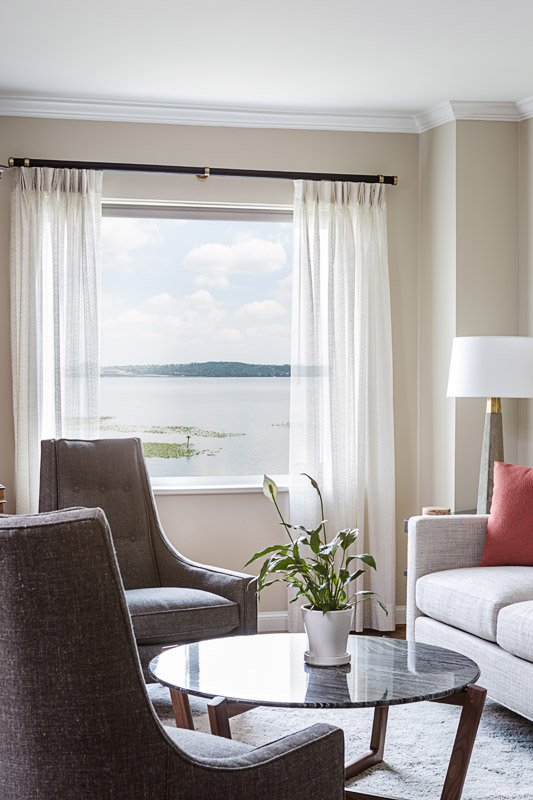 Living room with large window overlooking a lake, with sheer white curtains, a potted plant on a glass-top coffee table, a gray armchair, a white sofa with a red cushion, and a tall white lamp.
