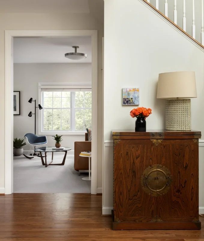 View of a living room through a doorway, with a wooden cabinet topped by a beige lampshade and a black vase with orange flowers in the foreground.