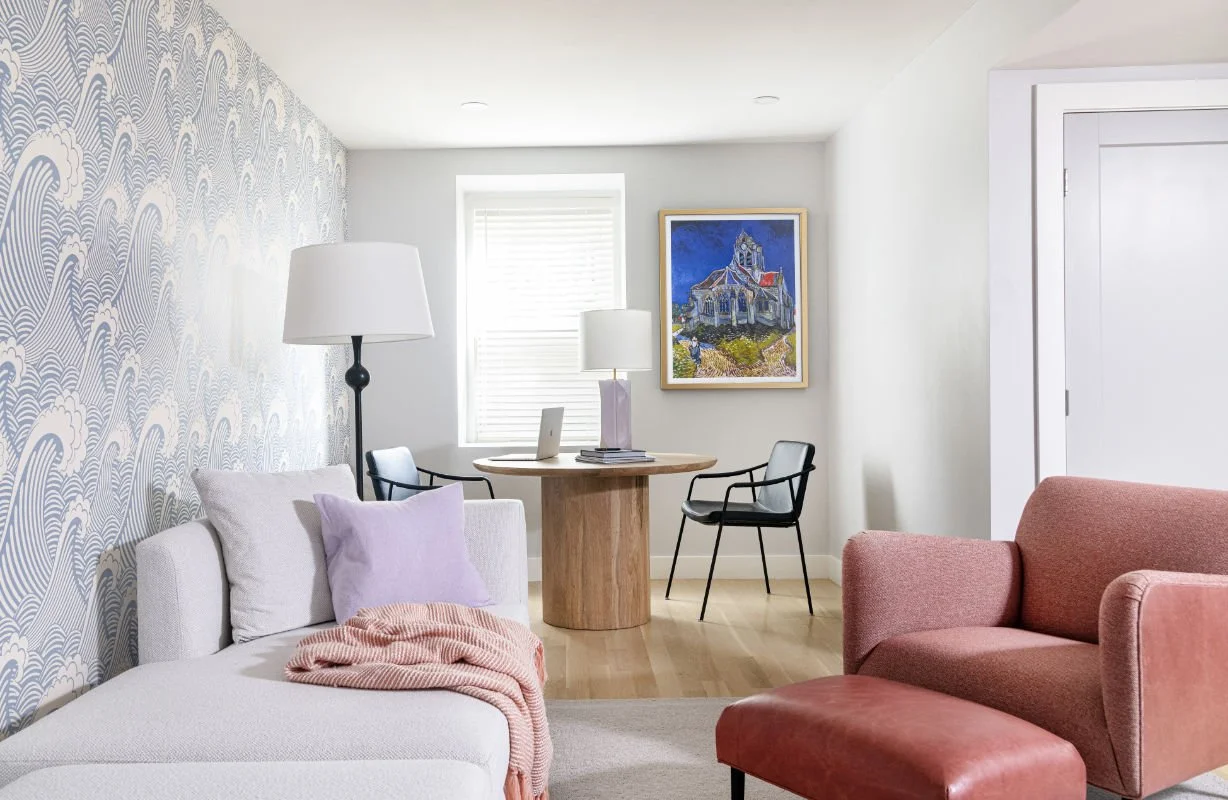 Living room with patterned accent wall, white sofa with pink blanket and lavender pillow, pink armchair with matching ottoman, round wooden table with two black chairs, standing lamp, window with blinds, and a painting of a house on the wall.