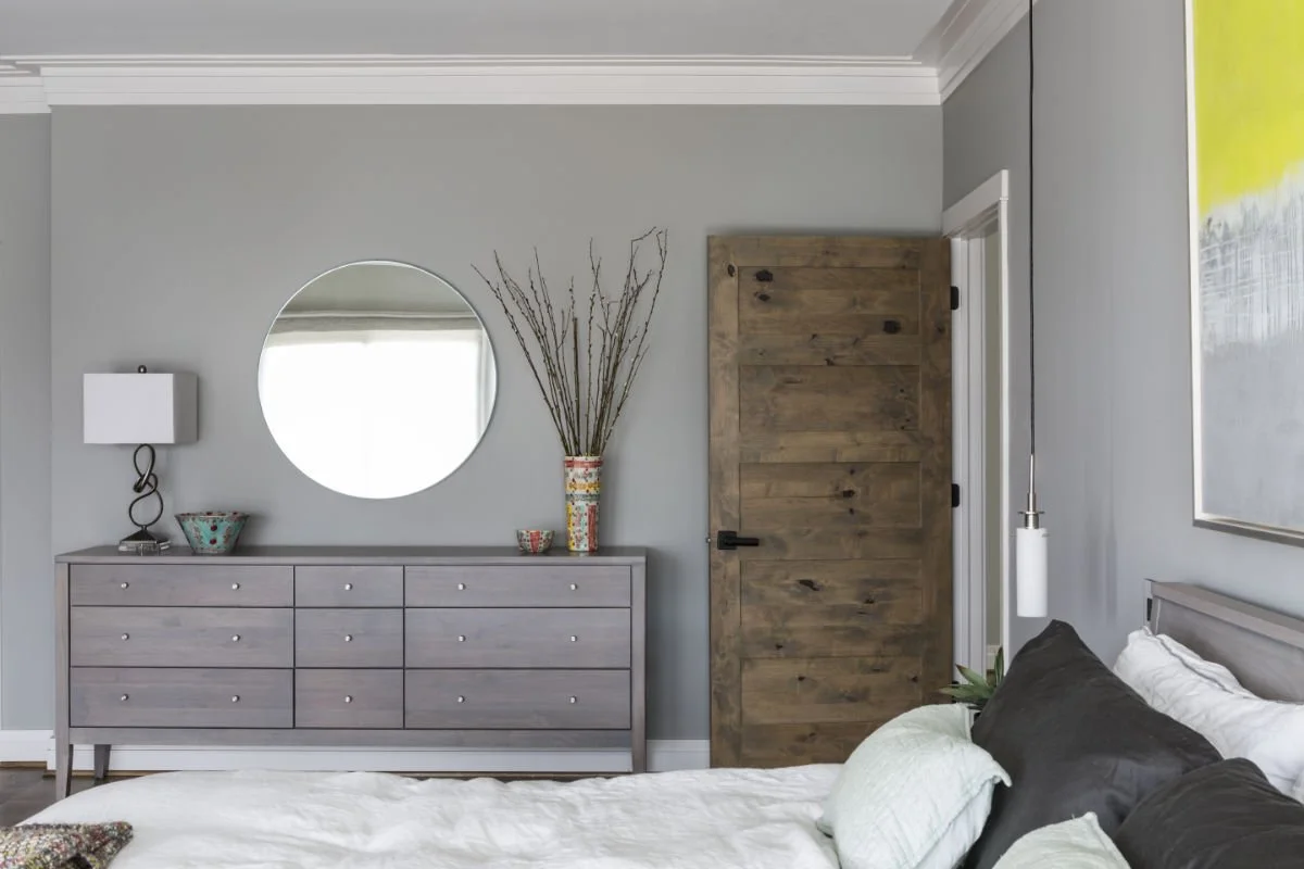 A bedroom with a gray dresser, a round mirror, a decorative vase with tall twigs, and a wooden door. Part of a bed with white and dark pillows is visible.