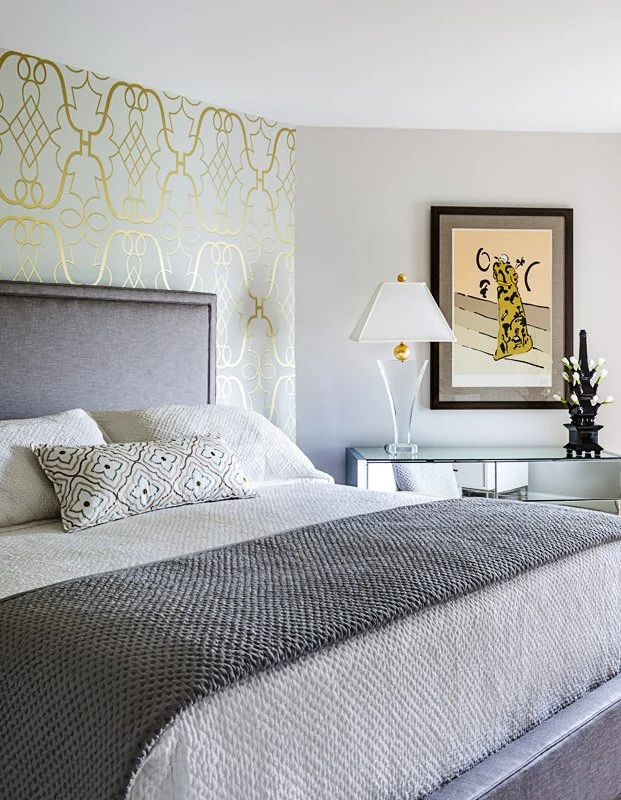 A bedroom with a bed featuring a gray headboard, white bedding, and decorative pillows. There is a gray nightstand with a white and gold lamp, framed artwork of a stylized animal, and a black decorative object with white flowers on a glass tabletop.