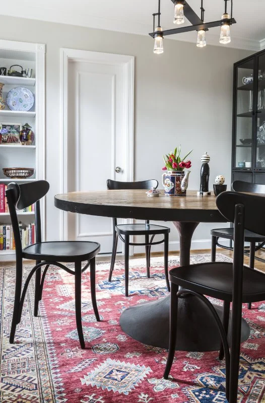 A dining room with a round wooden table surrounded by four black chairs, a patterned red and blue rug, a white door, and a black display cabinet filled with dishes and glassware. There is a flower arrangement and decorative items on the table, and a