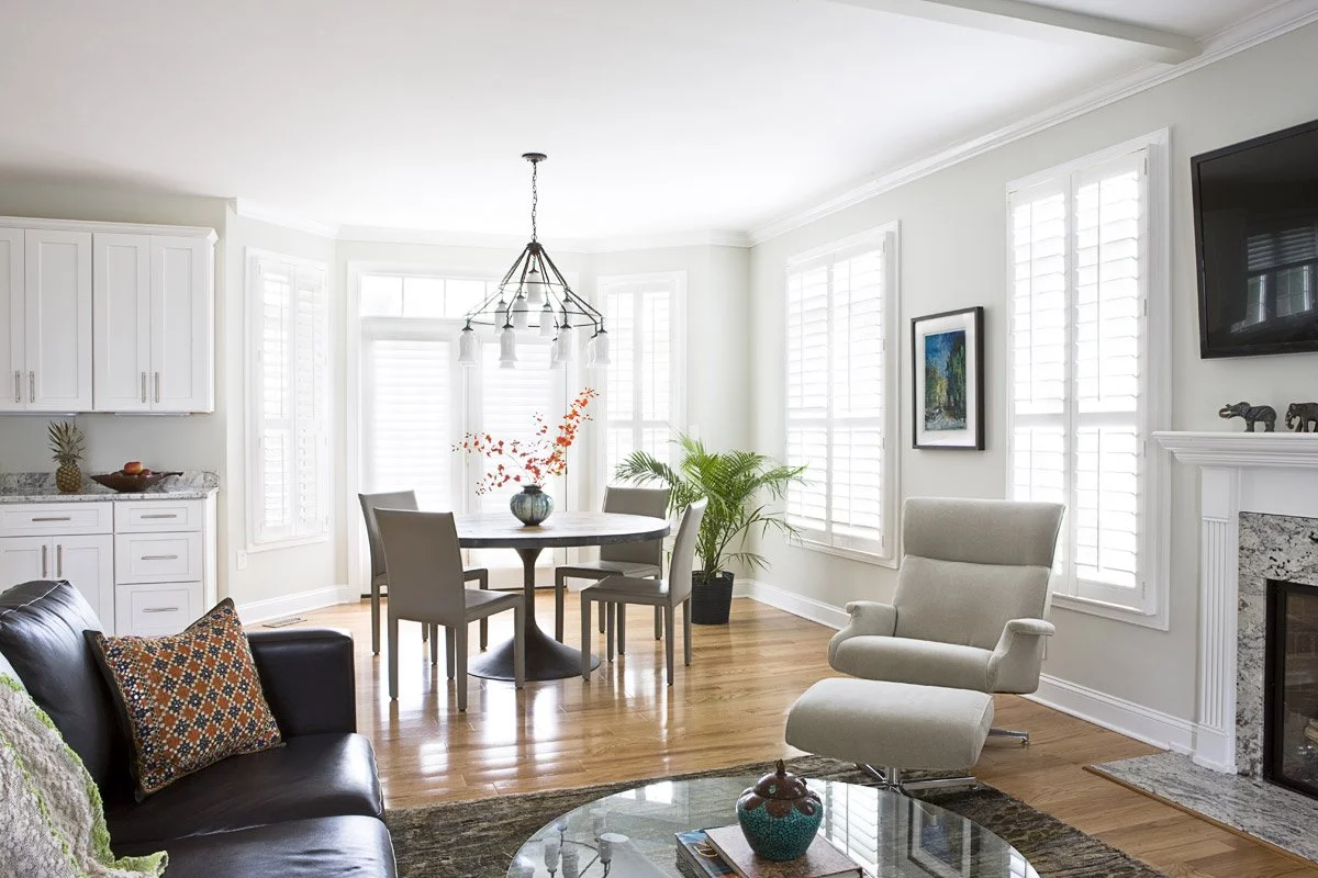 Bright living room with white walls, hardwood floors, a beige armchair and a black leather sofa, a glass coffee table, and a dining area with a round table and four chairs. Multiple windows with white plantation shutters and a fireplace with a mantel