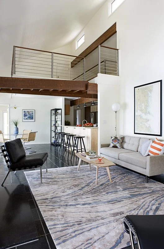 Open-concept living room and kitchen with high ceilings, large windows, modern furniture, and a staircase leading to an upper loft area.