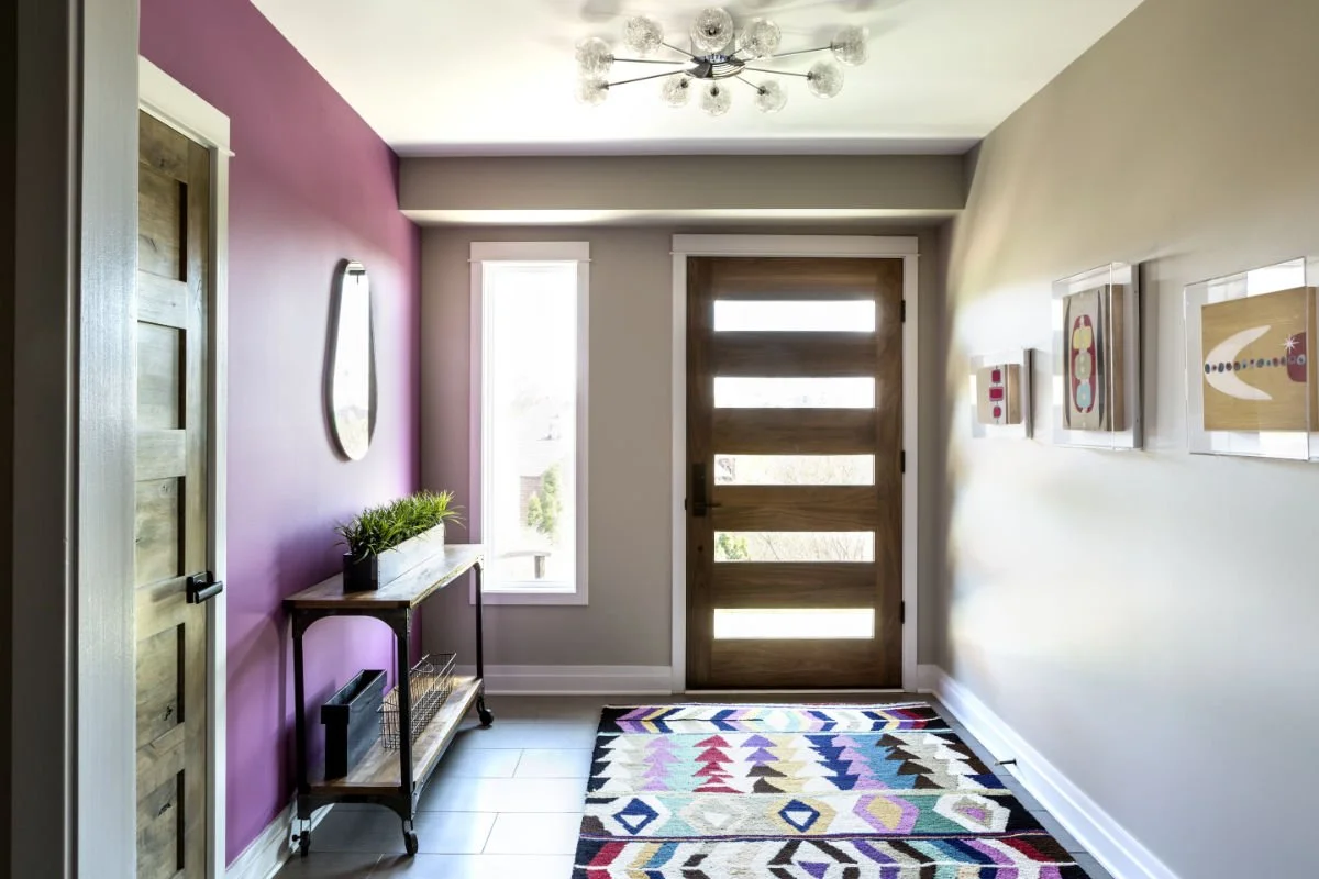 Modern entryway with a purple accent wall, a wooden front door with horizontal glass panels, a colorful geometric patterned rug, a narrow console table with green plants, a wall mirror, and framed artwork, illuminated by an overhead light fixture.