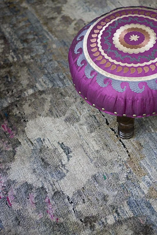 A round, purple upholstered stool with a colorful embroidered top sitting on a patterned rug.