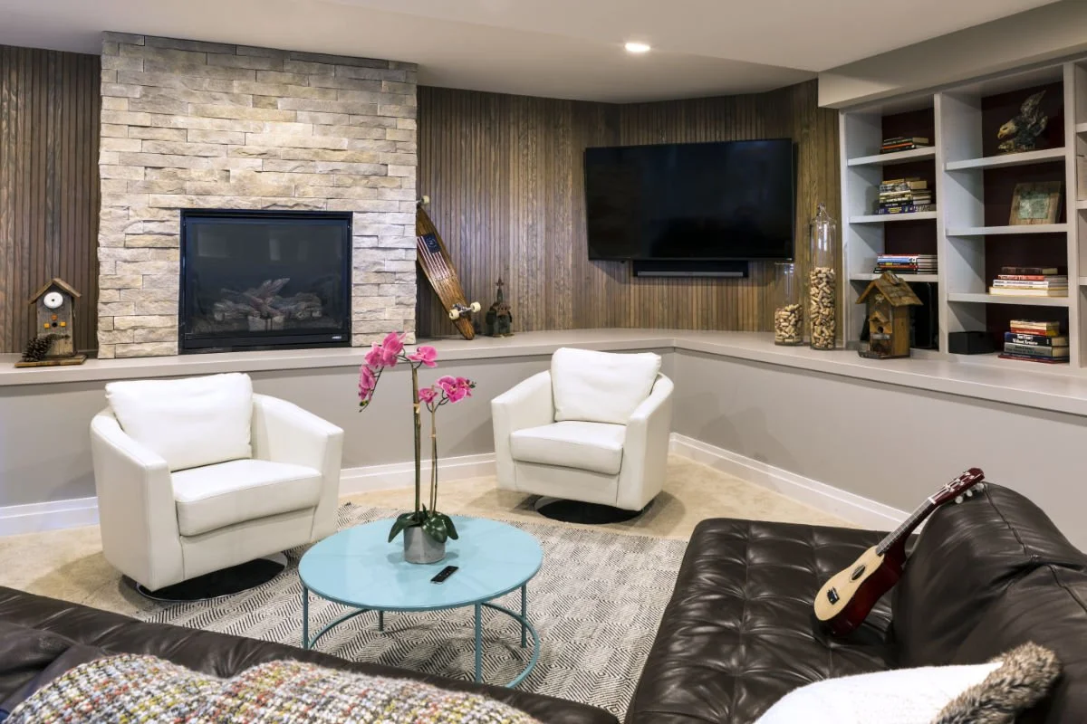 Living room with a fireplace, two white chairs, a black leather couch with an acoustic guitar, a blue coffee table with a pink orchid, built-in shelves with books, a wall-mounted flat-screen TV, and decorative objects.