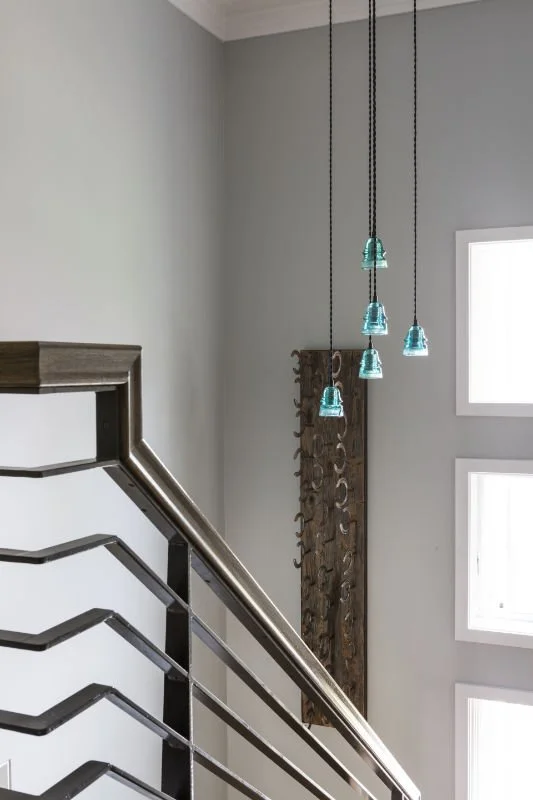 Interior view of a modern home staircase with black and wood railing, a decorative wooden wall hanging, and five hanging glass pendant lights in a light-colored room with large windows.