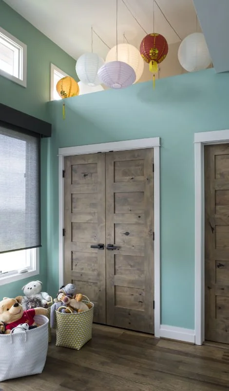 A corner of a room with light green walls, two wooden sliding closet doors, a window with a black blind, and a ceiling decorated with colorful paper lanterns. There are baskets filled with stuffed toys near the window.
