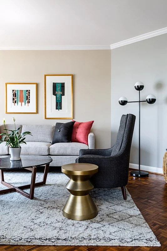 Living room with gray sofa, colorful pillows, modern black armchair, potted plant, abstract art on the wall, gold side table, black glass coffee table, and tripod floor lamp with globe lights, hardwood floor, and light-colored walls.