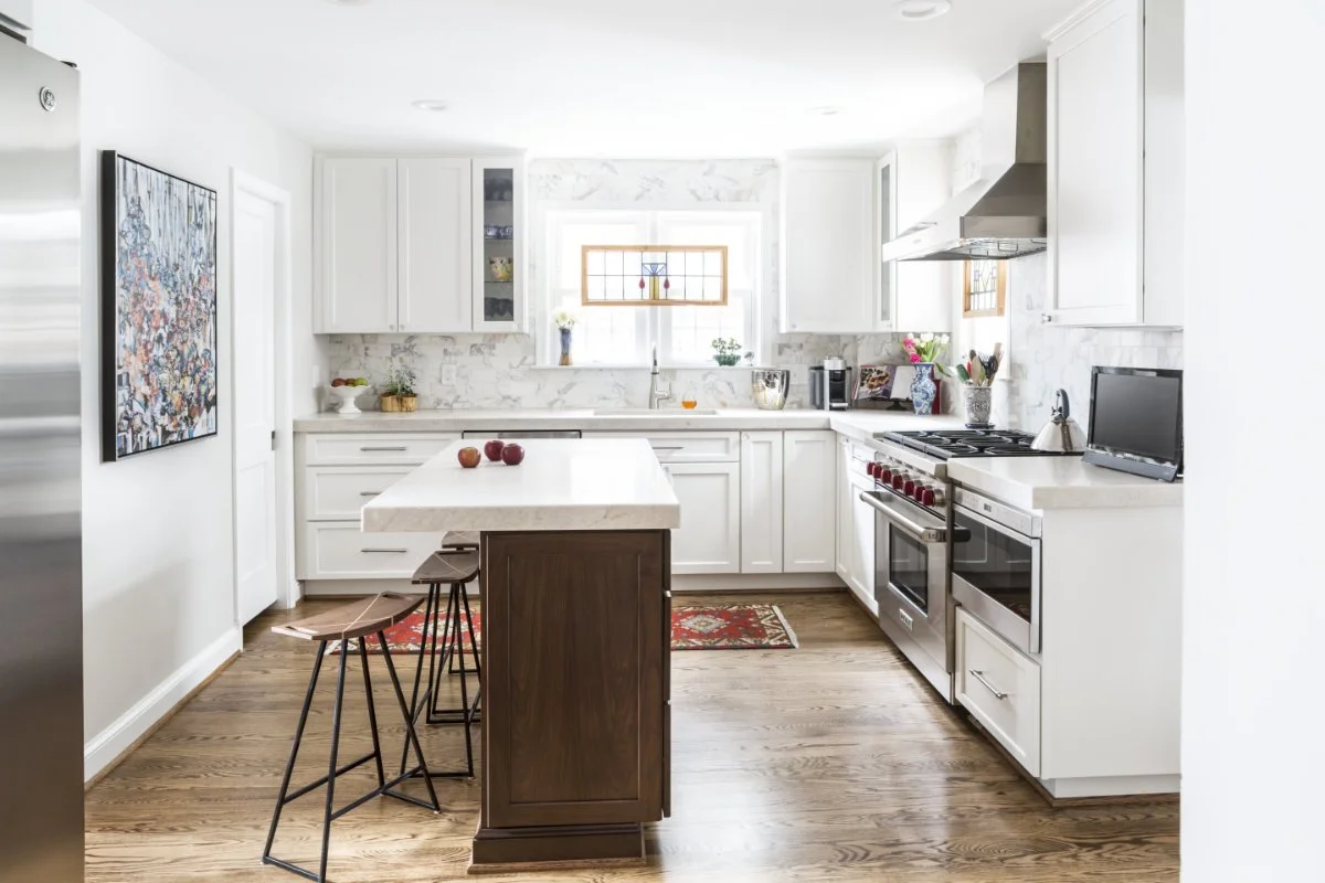 Bright white kitchen with wooden flooring, white cabinets, stainless steel appliances, a small vase with flowers on the window sill, and a central island with chairs. Apples are on the island, and a colorful rug is on the floor.