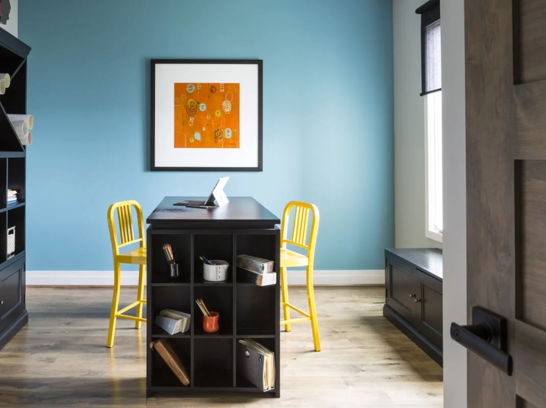 Home office with a black bookshelf, two yellow chairs, a blue wall, a framed orange abstract art, a window with a black blind, and hardwood floors.
