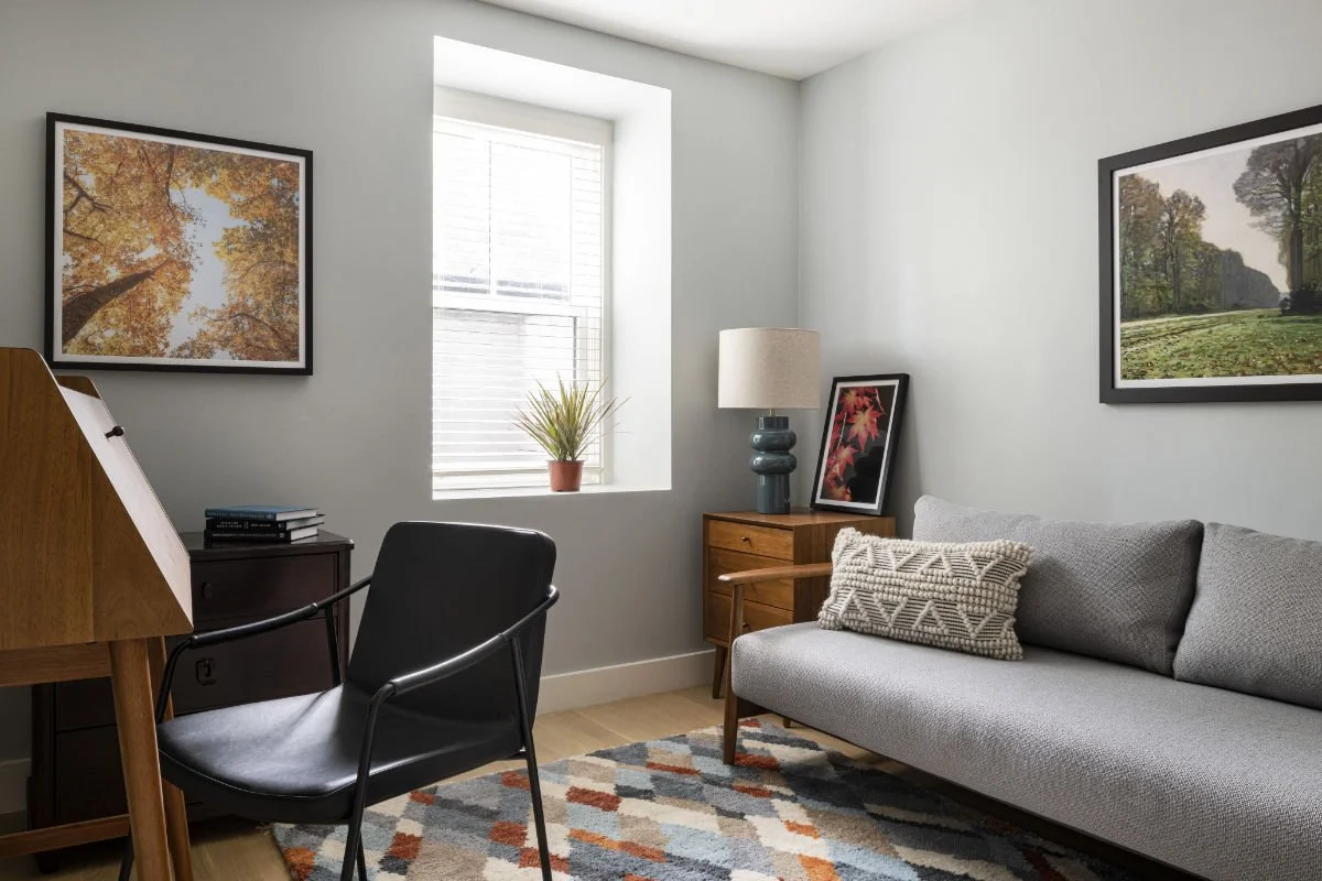 Living room with a gray sofa, a black armchair, wooden side table with a lamp, framed pictures on the walls, a window with blinds, and a patterned rug.