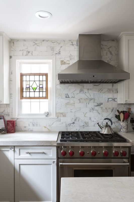 Modern kitchen with white cabinetry, marble backsplash, stainless steel range hood, stainless steel stove, small window with stained glass, and kitchen utensils.