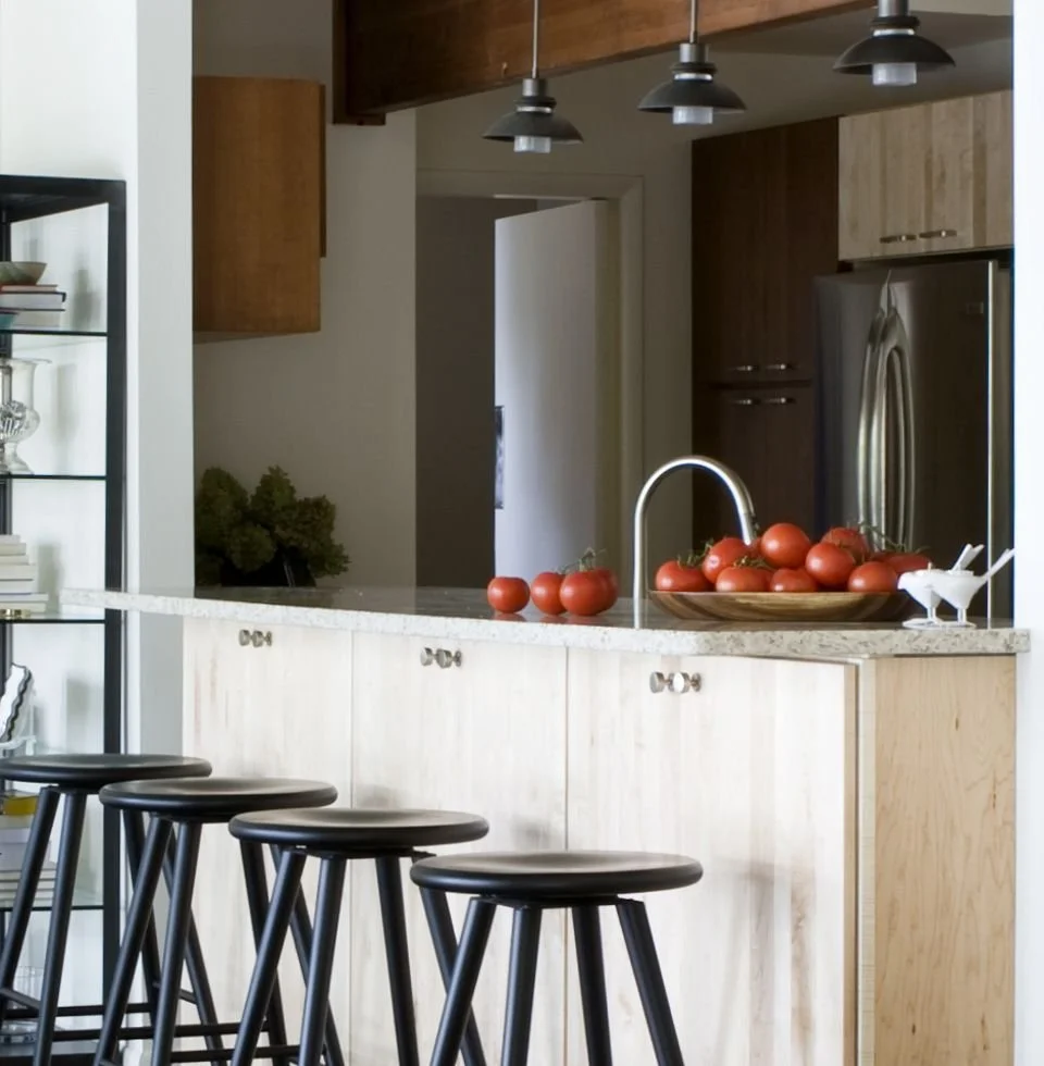 Kitchen with a light-colored island counter, black bar stools, a bowl of fresh tomatoes, and modern black pendant lights.