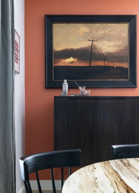 Interior of a dining room with a round marble table, black chairs, a black cabinet, framed landscape painting of sunset and power lines on a reddish-orange wall, and a tray with bottles and glasses.