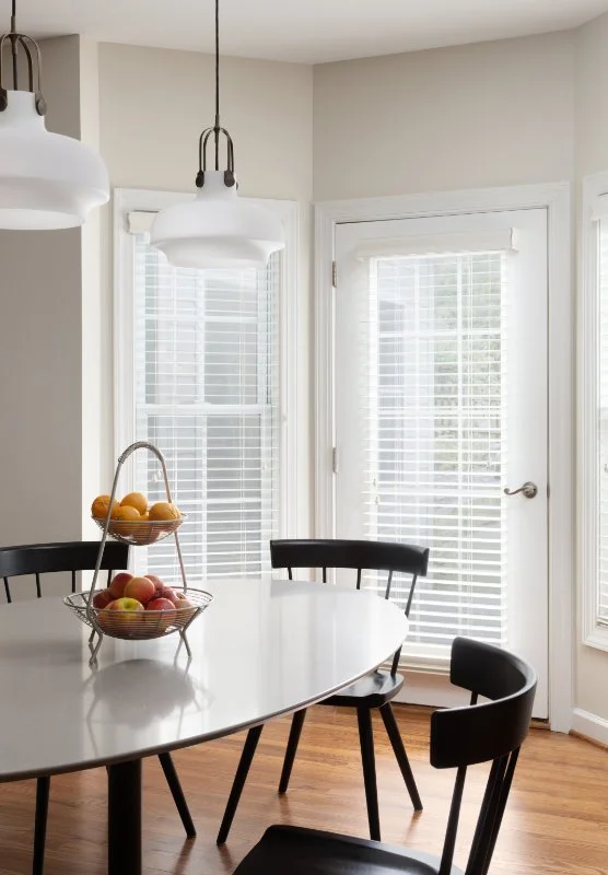 A bright kitchen corner featuring a white oval dining table with black chairs. On the table, there is a two-tier fruit basket filled with oranges and apples. The area is illuminated by two white pendant lights hanging from the ceiling, and the backgr