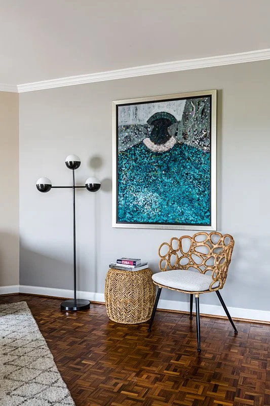 A living room corner with a floor lamp, a small woven side table with magazines, a modern rattan chair with a cushion, a framed mosaic art piece on the wall, and hardwood flooring.