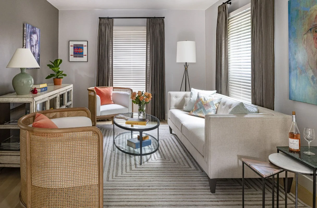 A modern living room with a white sofa, beige armchair, rattan chairs, and a glass coffee table with books and flowers. The room has gray walls, striped rug, and windows with white blinds and gray curtains. There are decorative paintings, a white con