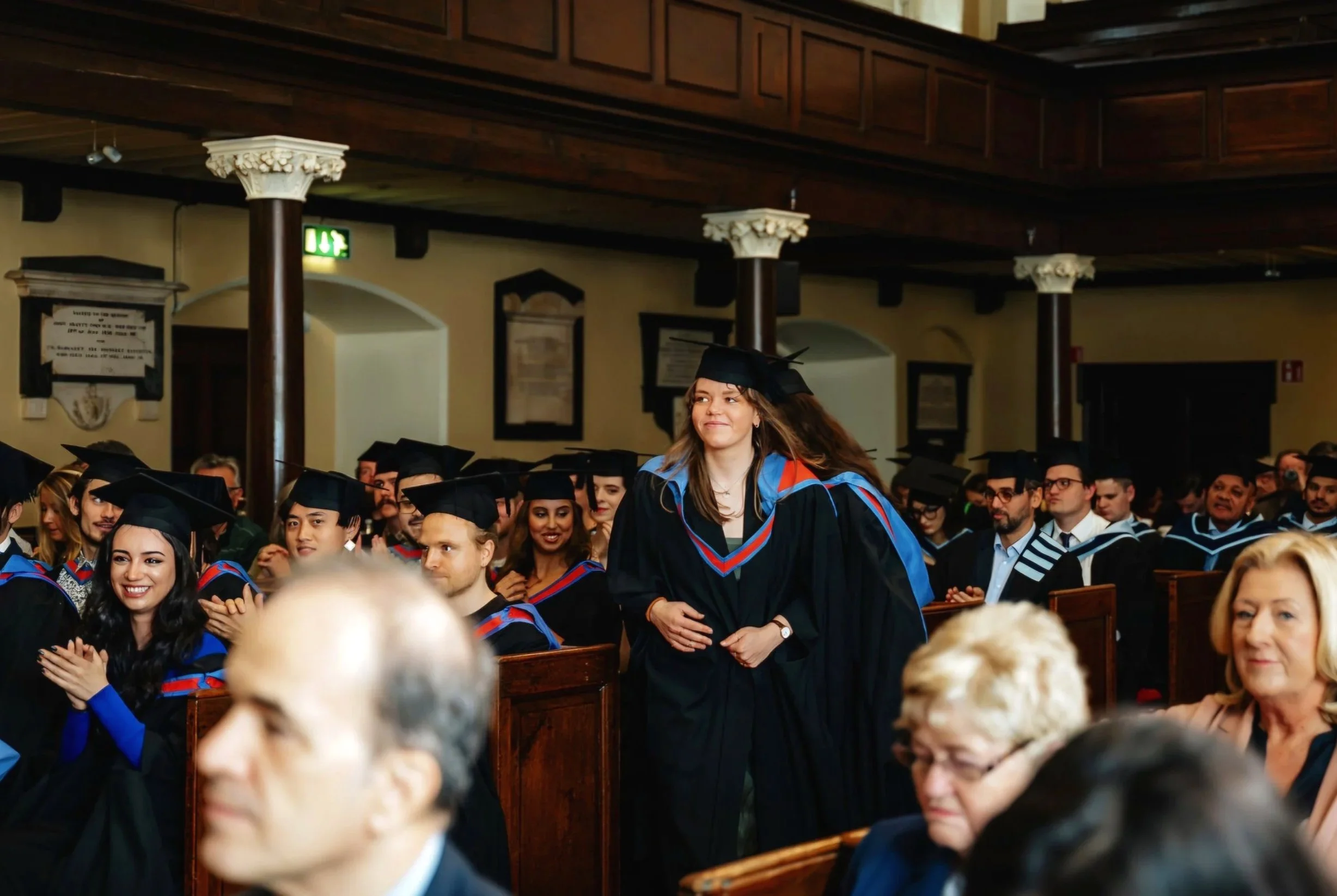 A group of graduates wearing caps and gowns at a graduation ceremony, sitting in rows inside a hall with wooden beams and columns, some are clapping and smiling.