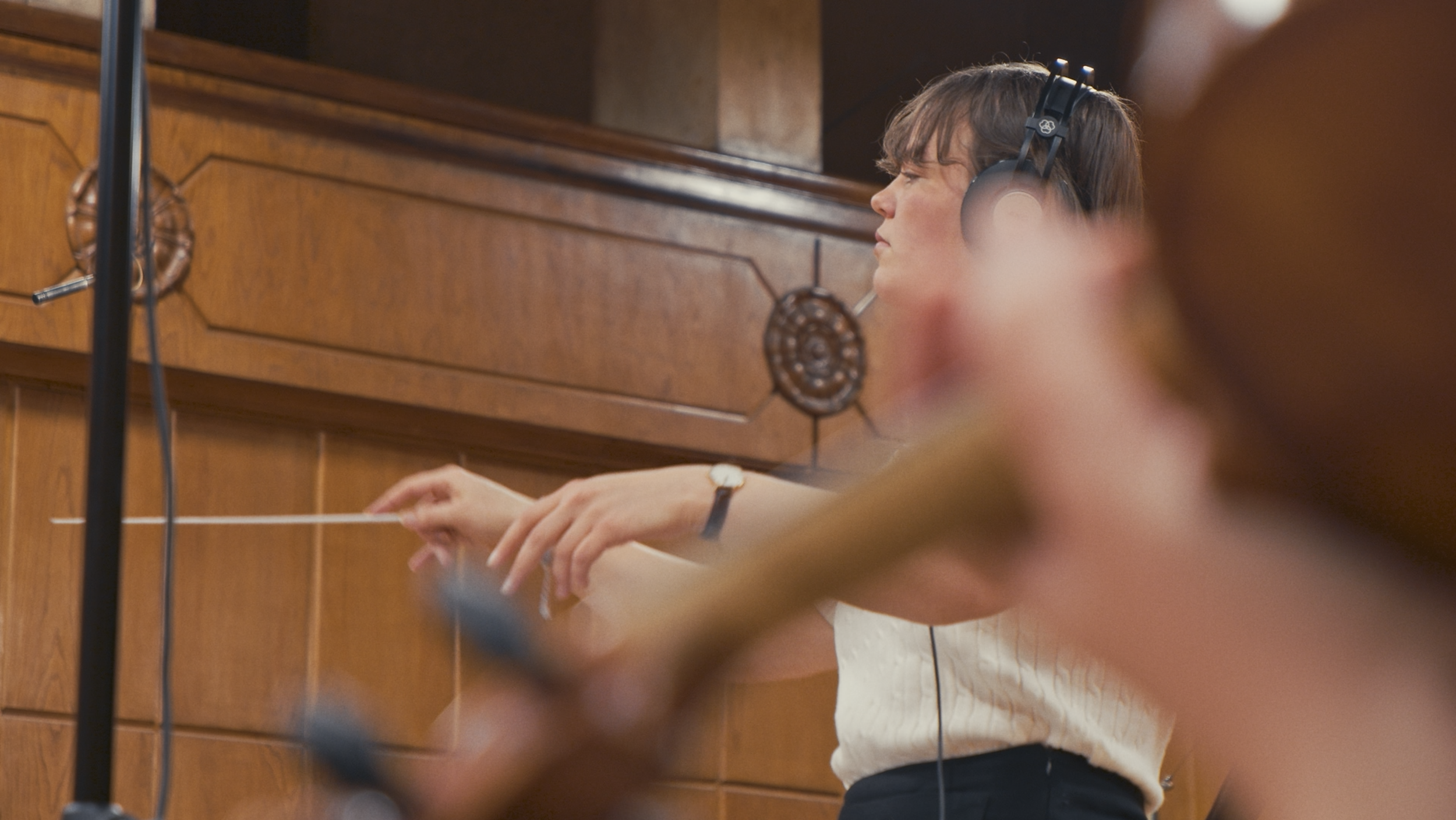 A woman in a music recording studio is wearing headphones and conducting an orchestra or musical ensemble, with her eyes closed and her hands raised.