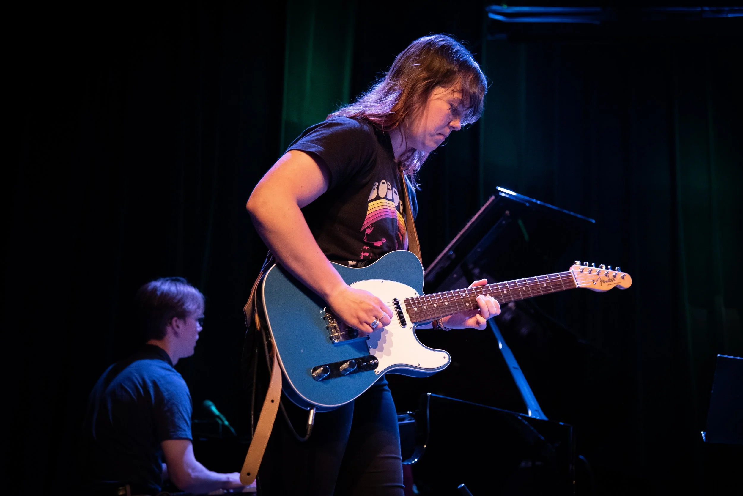 A woman playing an electric guitar on stage with a keyboardist in the background.