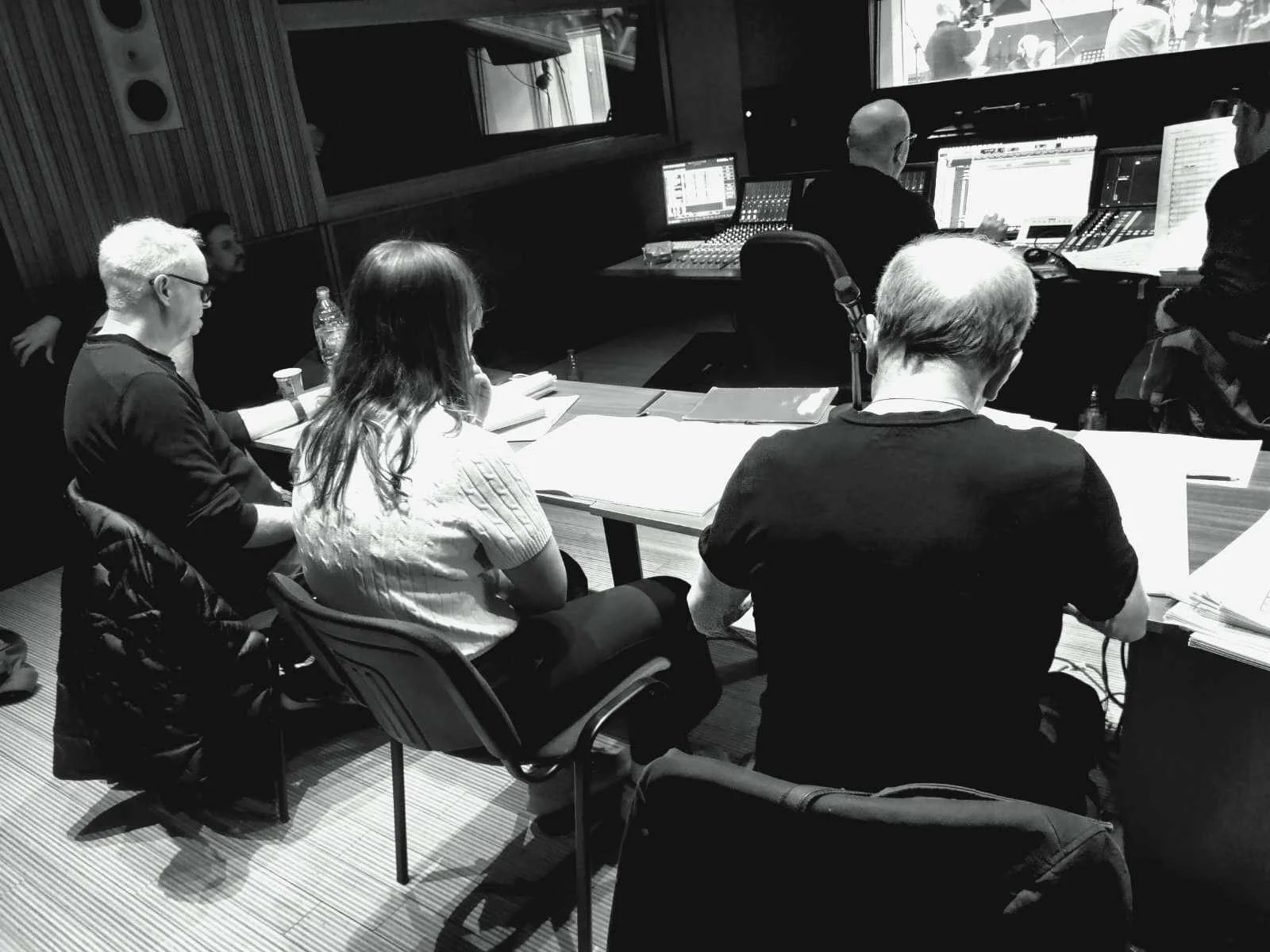A black and white photo of a recording studio with five people seated around a table, some taking notes or reading scripts, and four individuals working at mixing consoles and computers in the background.