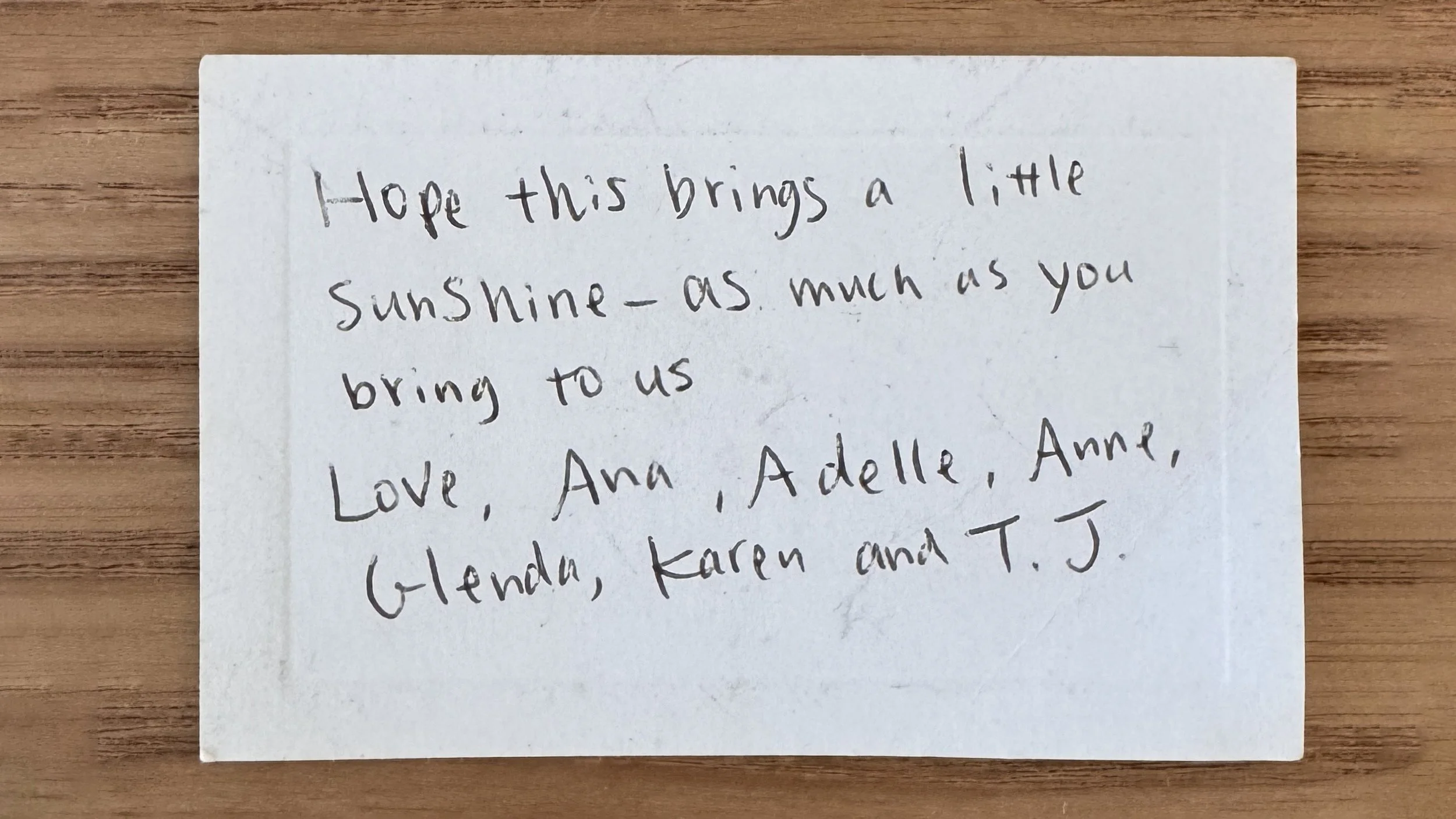 Handwritten note on white paper placed on a wooden surface with a message of encouragement that reads, “Hope this brings a little sunshine, as much as you bring to us. Love, Ana, Adelle, Anne, Glenda, Karen and T.J.”