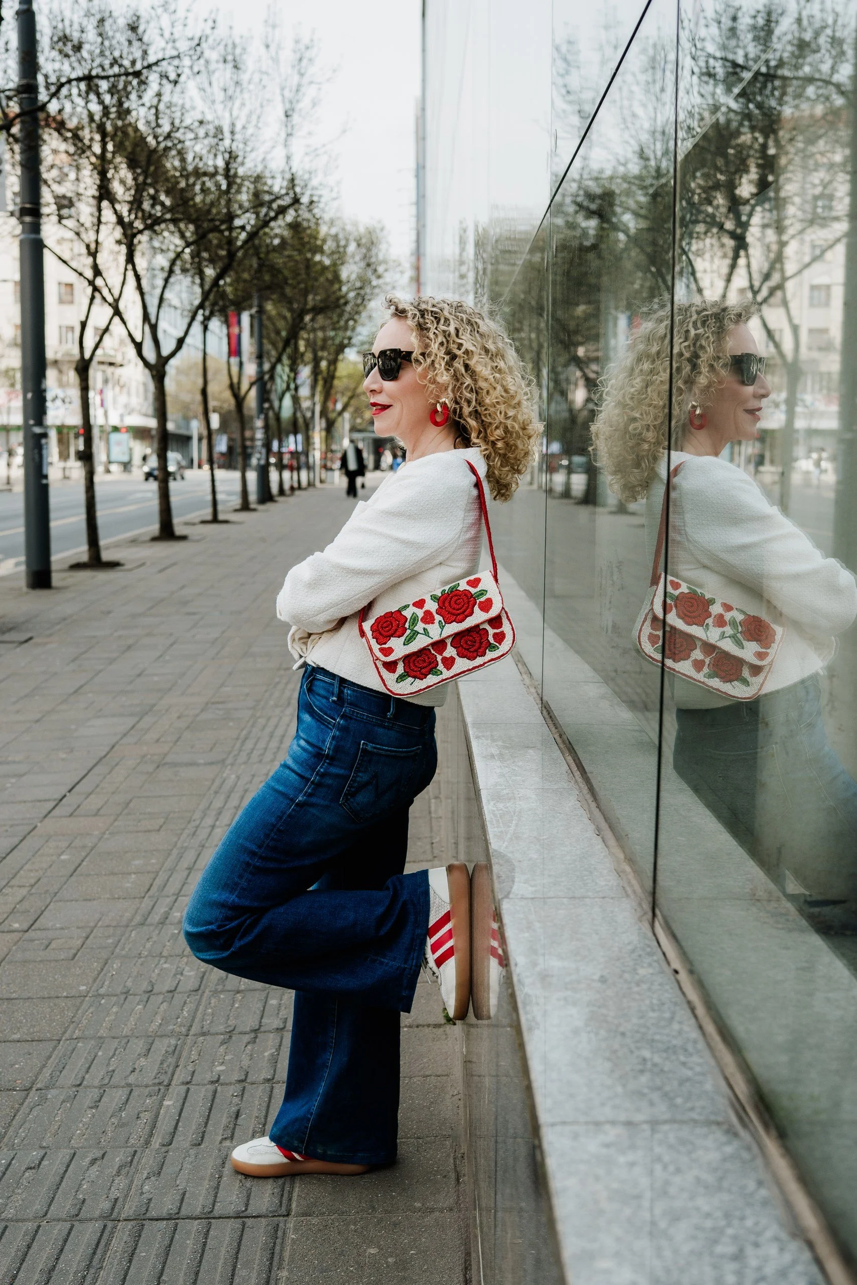 Mila Apostolovic, menopause coach, wearing a white sweater and jeans, standing next to a glass wall with her reflection visible.