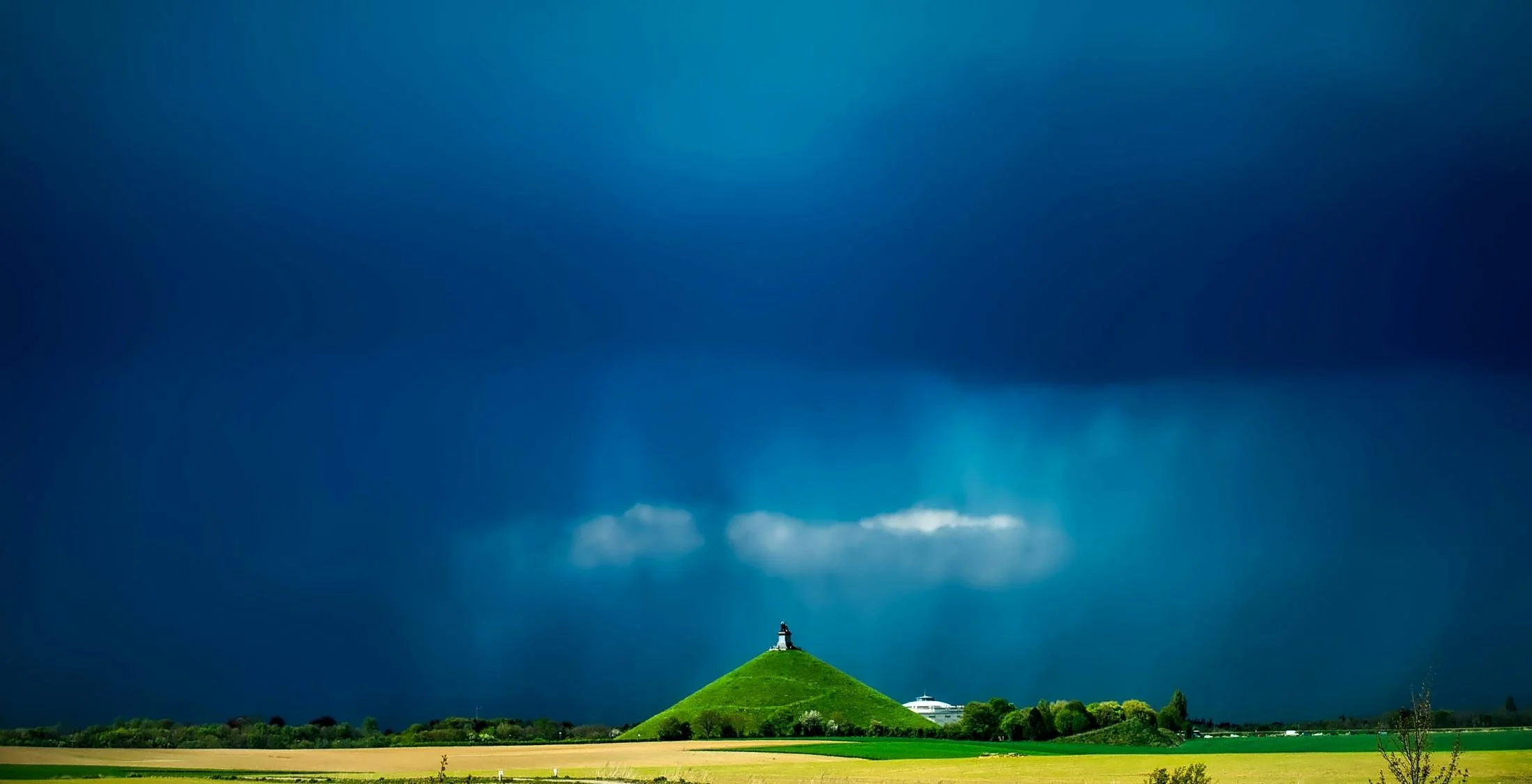 Paysage rural avec un mont verdoyant au centre, une statue ou un monument au sommet, sous un ciel dramatique avec des nuages sombres.