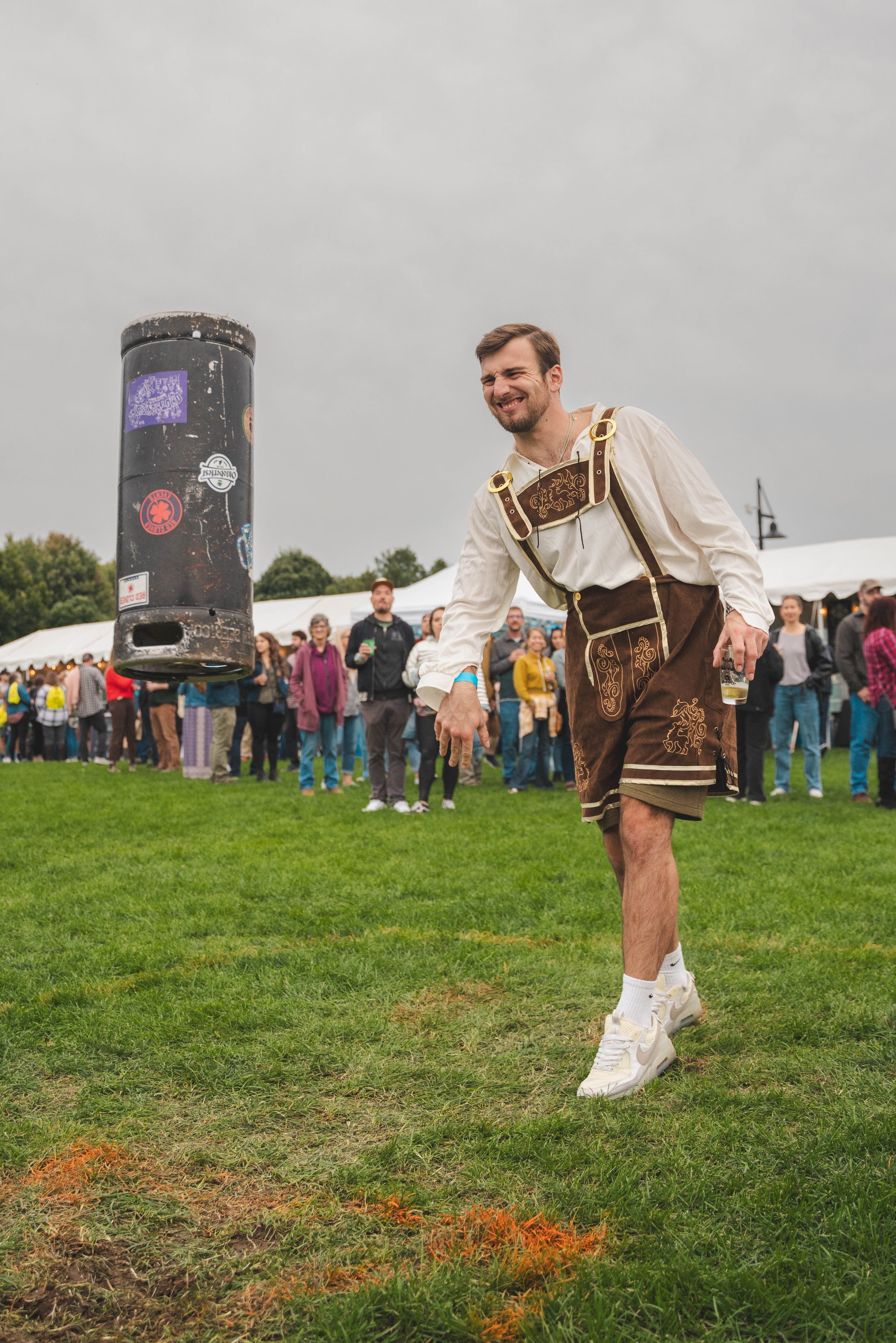 A man in traditional Bavarian attire tossing a beer stein in the air at an outdoor event, with a crowd of people in the background under cloudy skies.