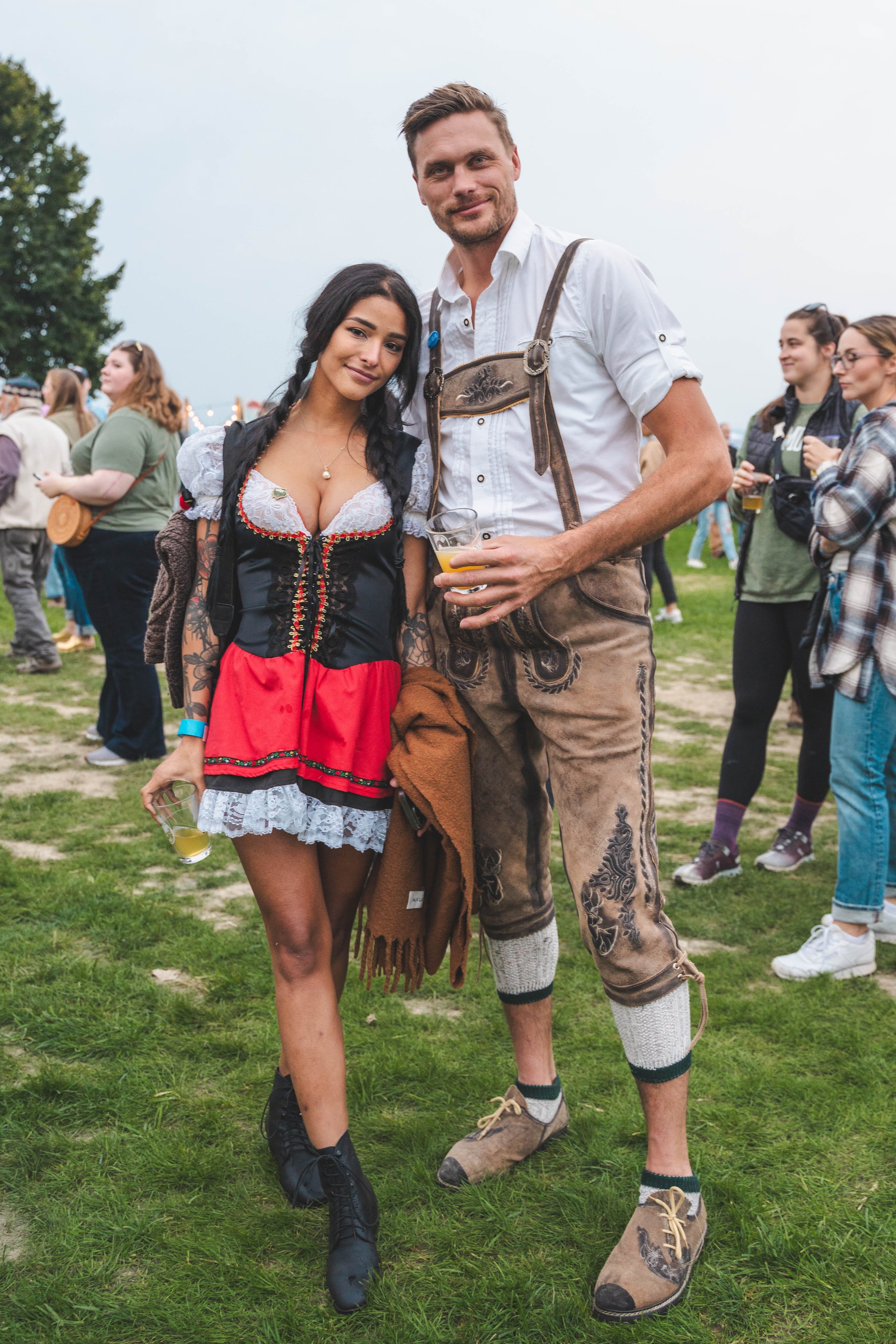 A man and woman dressed in traditional German attire standing outdoors at a festival, both holding drinks, with festival-goers in the background.