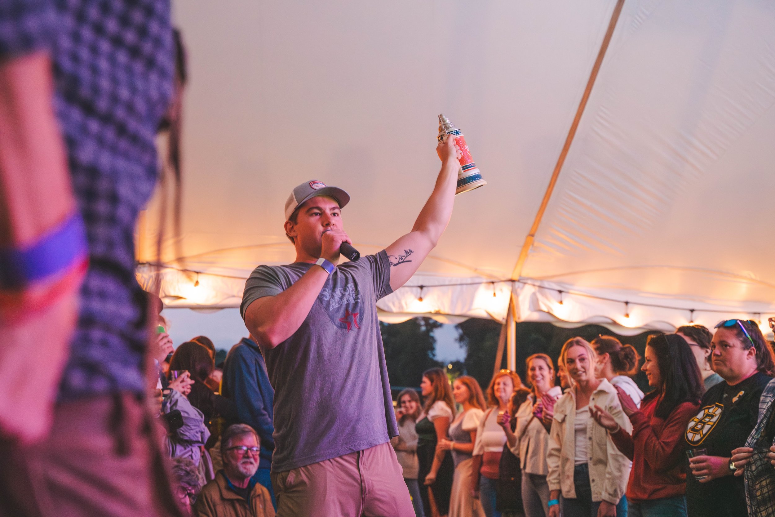 A man standing inside a large tent, holding a beer stein in one hand and a microphone in the other, speaking or singing to a crowd of diverse people.