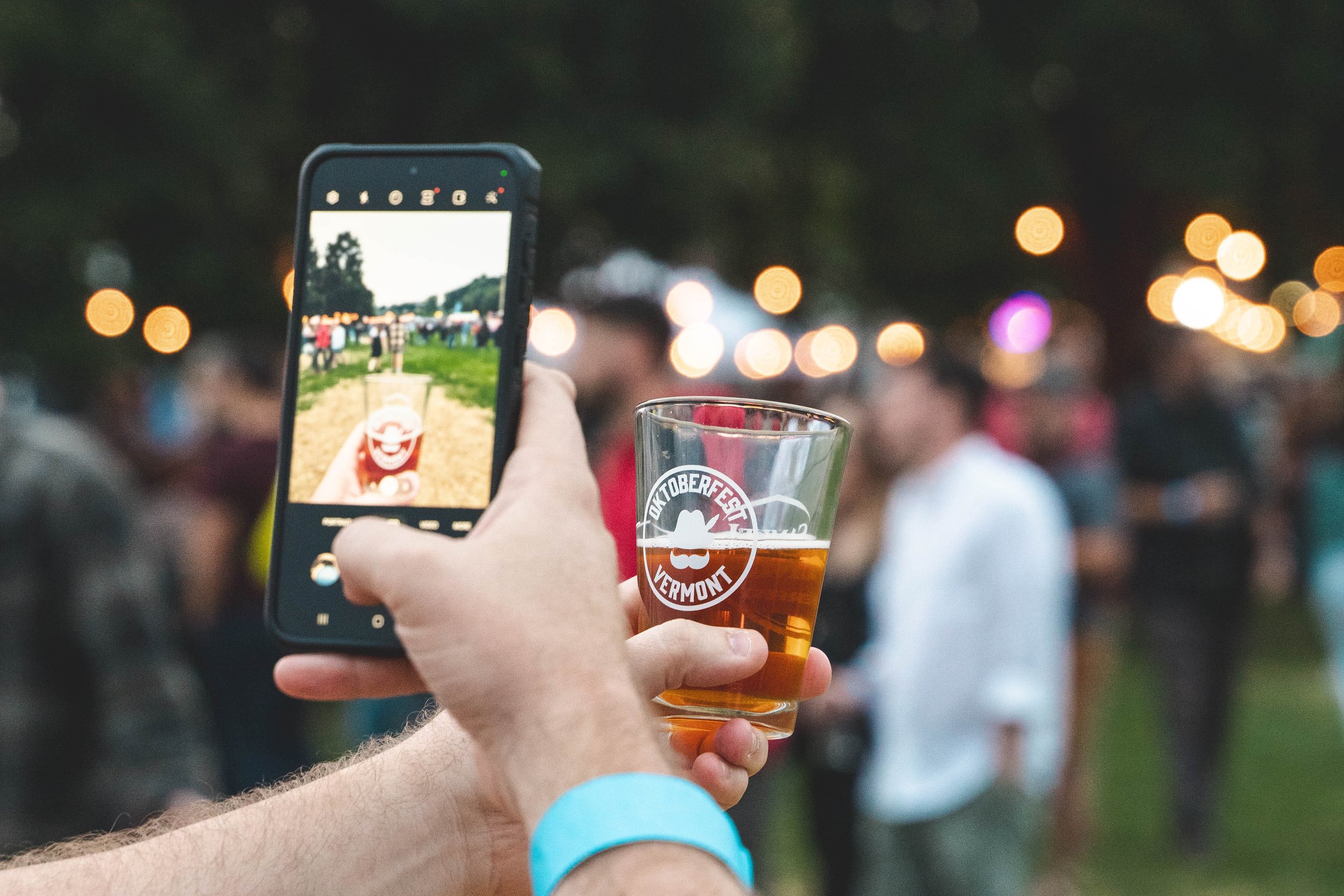 Person holding a smartphone and a glass of beer at a crowded outdoor festival during dusk, with blurred people and string lights in the background.