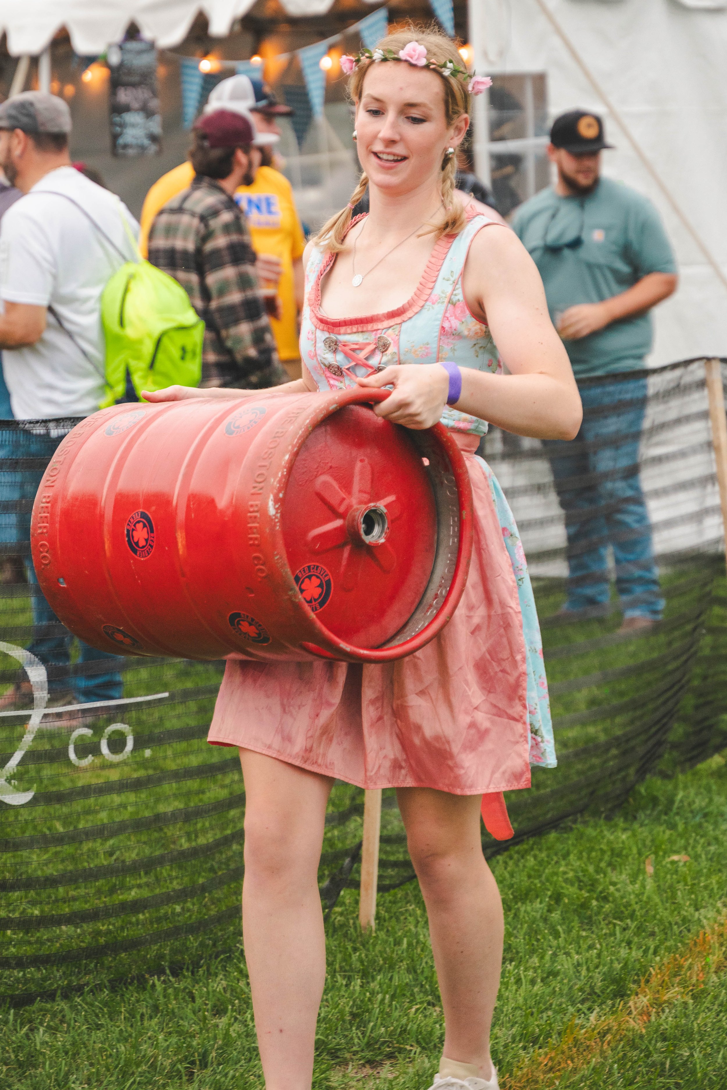 A young woman dressed in a traditional dirndl holding a red beer keg at an outdoor festival, with other people in casual attire in the background.