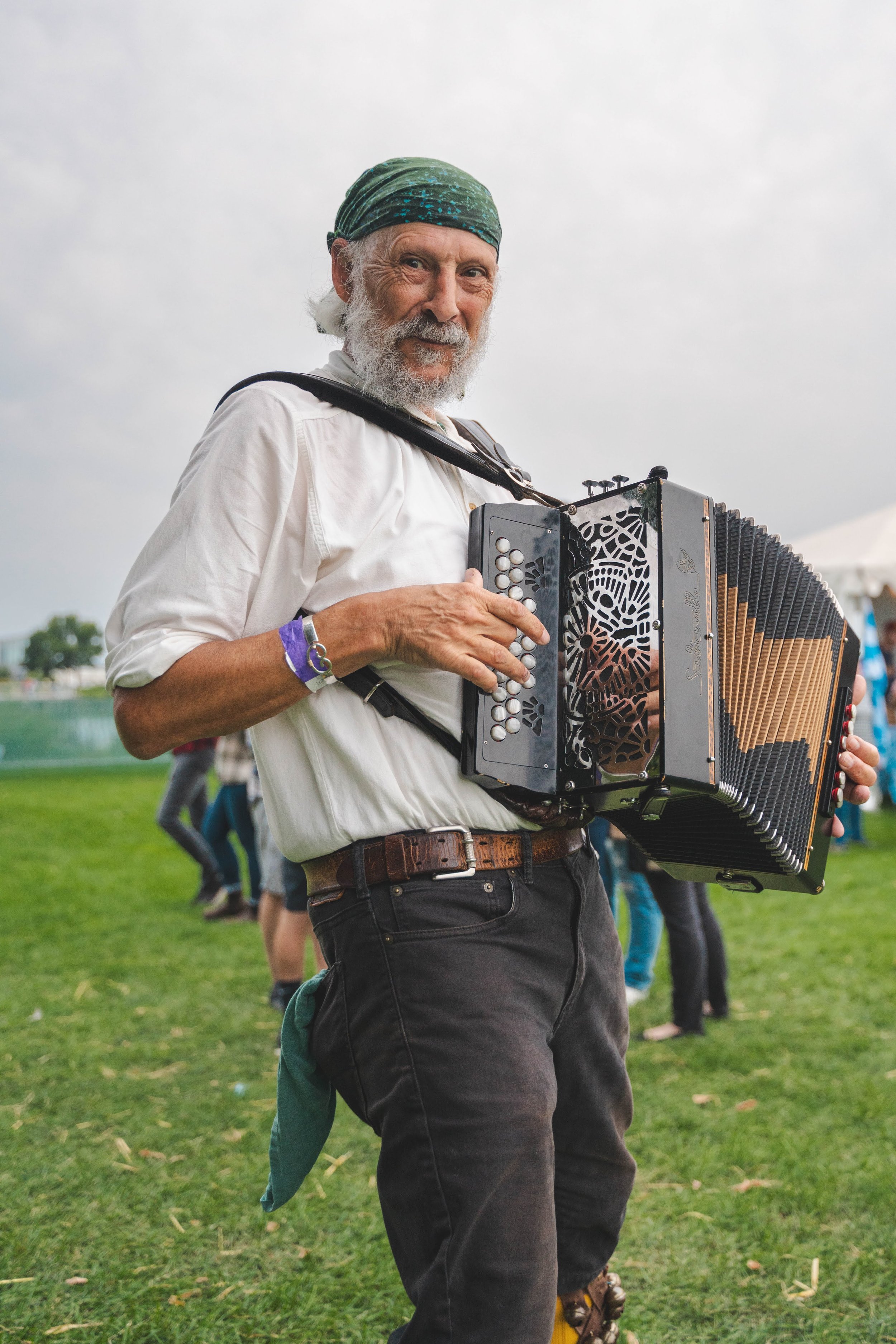 An older man with a gray beard and a green bandana playing a button accordion outdoors at a festival. He is wearing a white shirt, black pants, and a brown belt, with people and tents in the background.