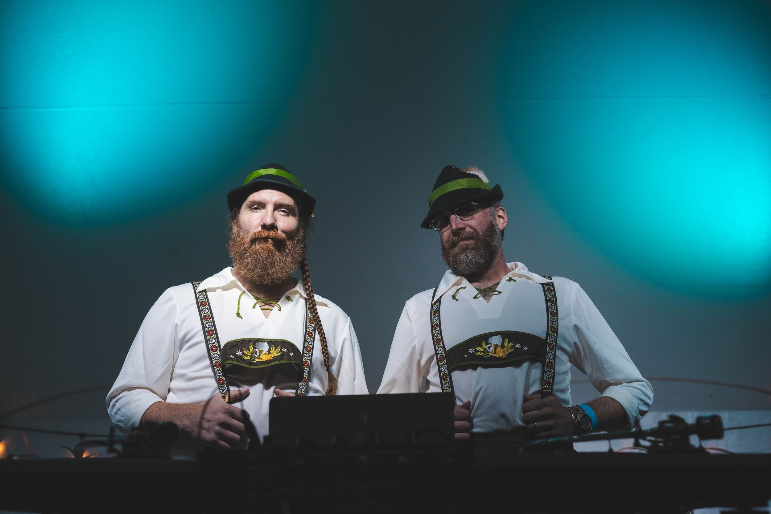 Two men standing behind a DJ setup, wearing traditional German attire with lederhosen and hats, under blue lighting.