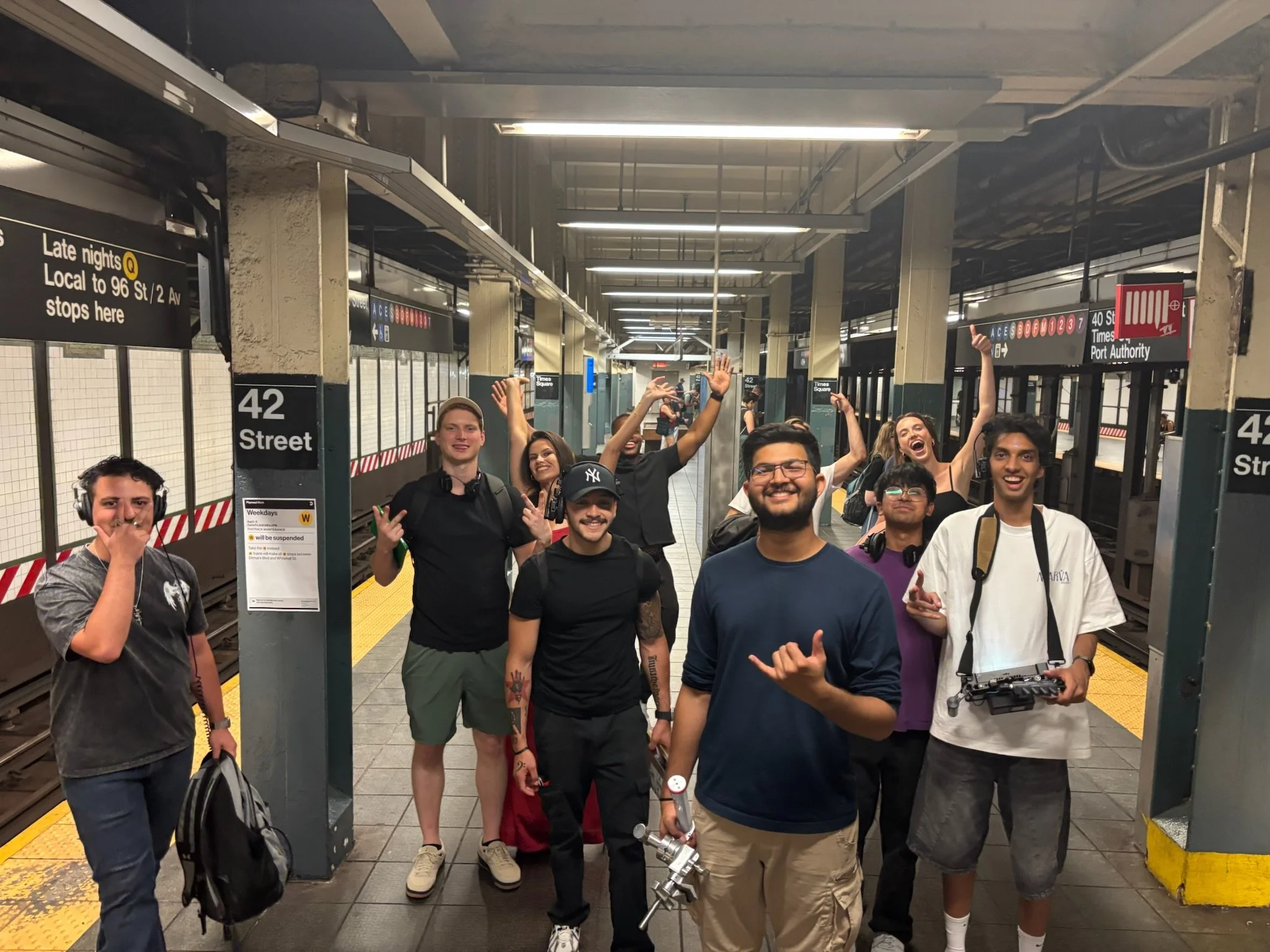 Cast and Crew of Another Day in New York standing on a subway platform at 42nd Street, smiling and waving at the camera, with some holding cameras and wearing backpacks.