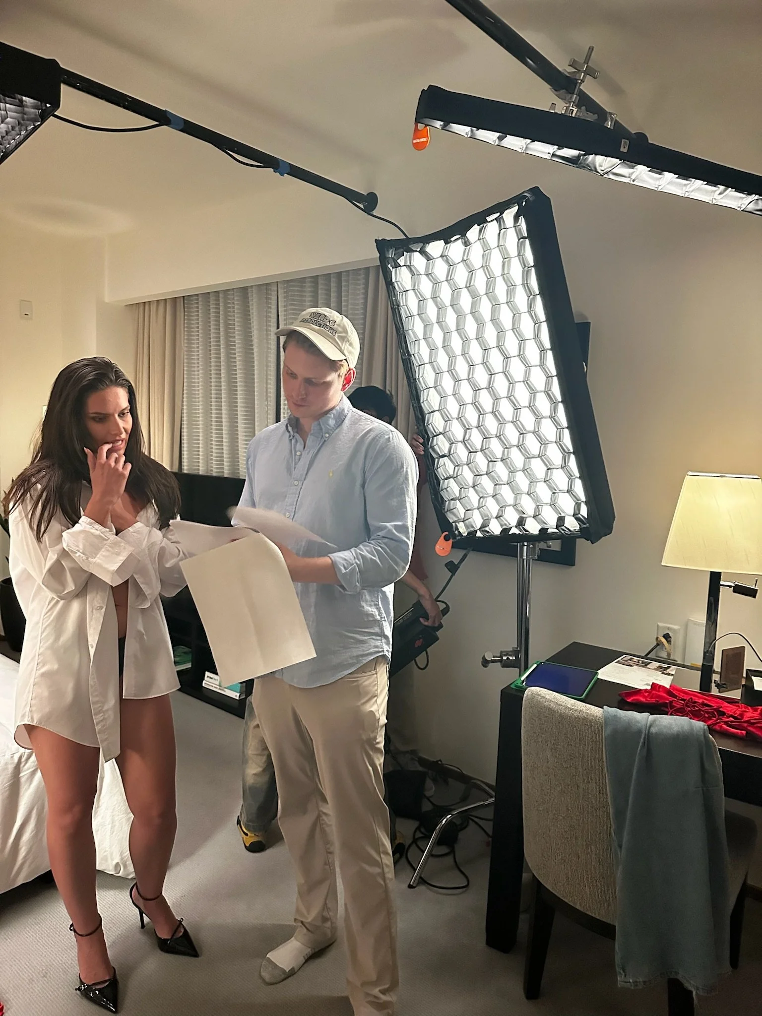 A woman in a white shirt and black heels, and a man in a light blue shirt and beige pants, are looking at papers in a room with professional lighting equipment, a lamp, and a gray armchair.