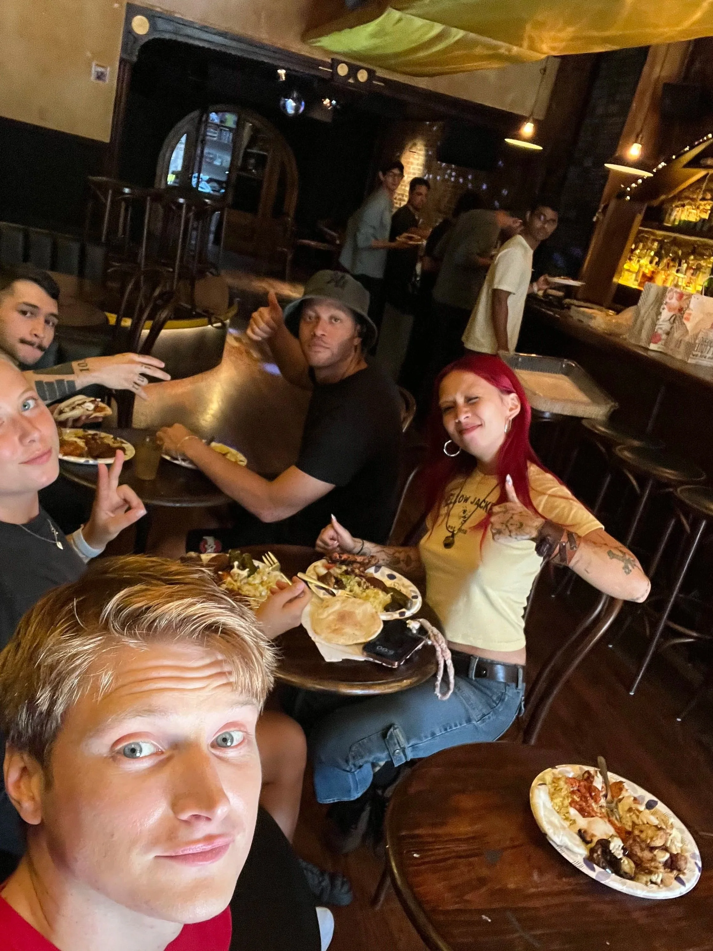 People sitting around a wooden table in a restaurant, with pizza on plates. The photo is a selfie taken by a person with light-colored hair and facial tan, in the foreground. Four other people are visible in the background, some looking at the camera