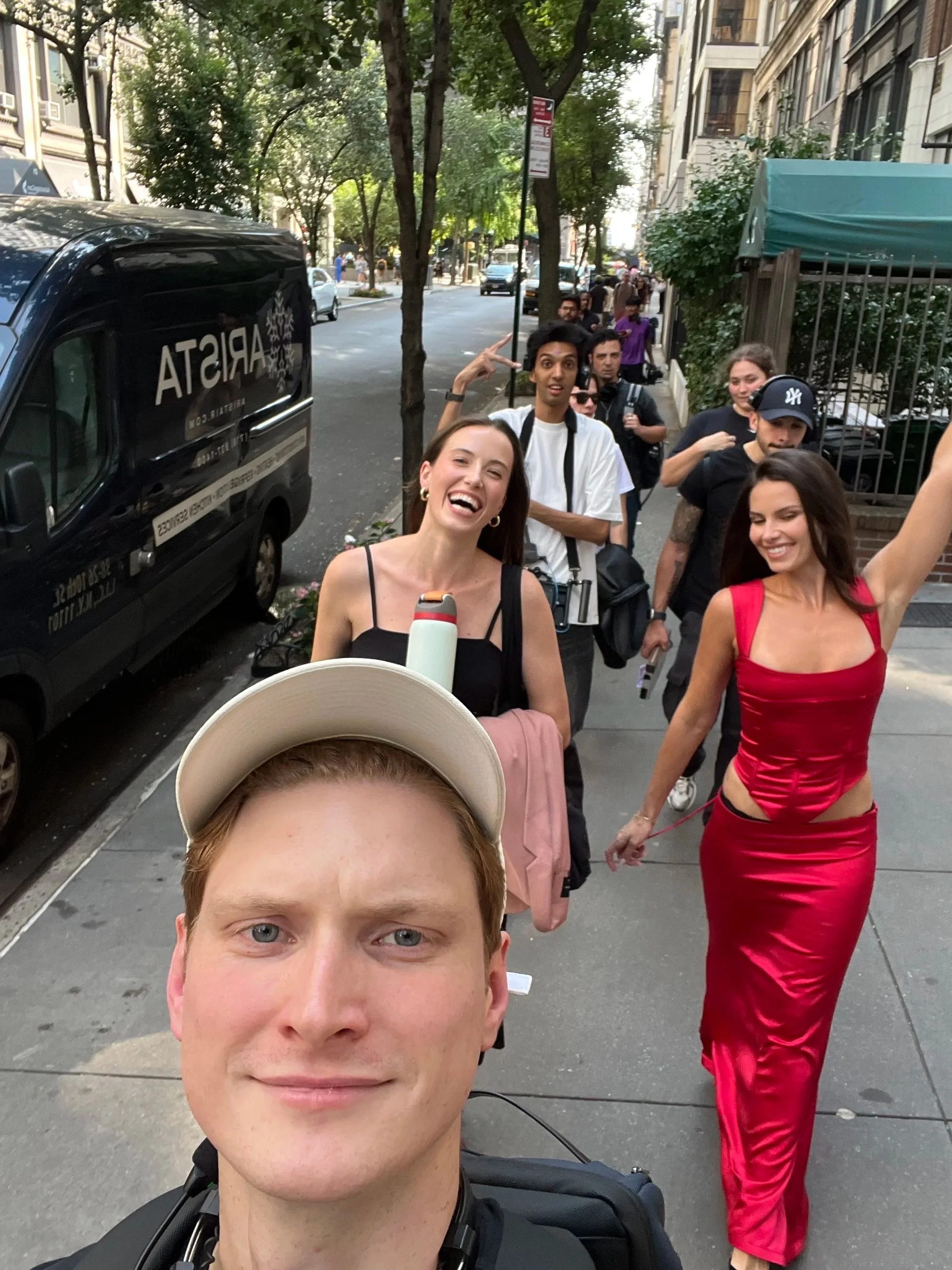 A group of people walking on a city sidewalk, with some smiling and taking selfies, including a woman in a red dress at the front.