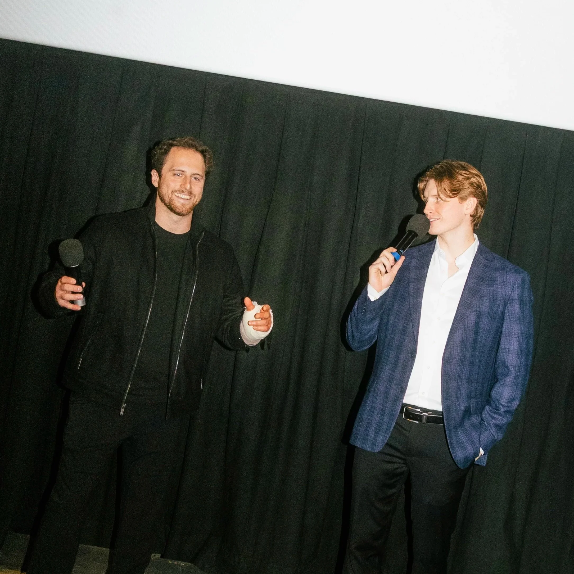 Adam and Sean Pine standing on stage at the NYC premier for their film Another Day in New York, one with a microphone, background features a black curtain and a white ceiling.