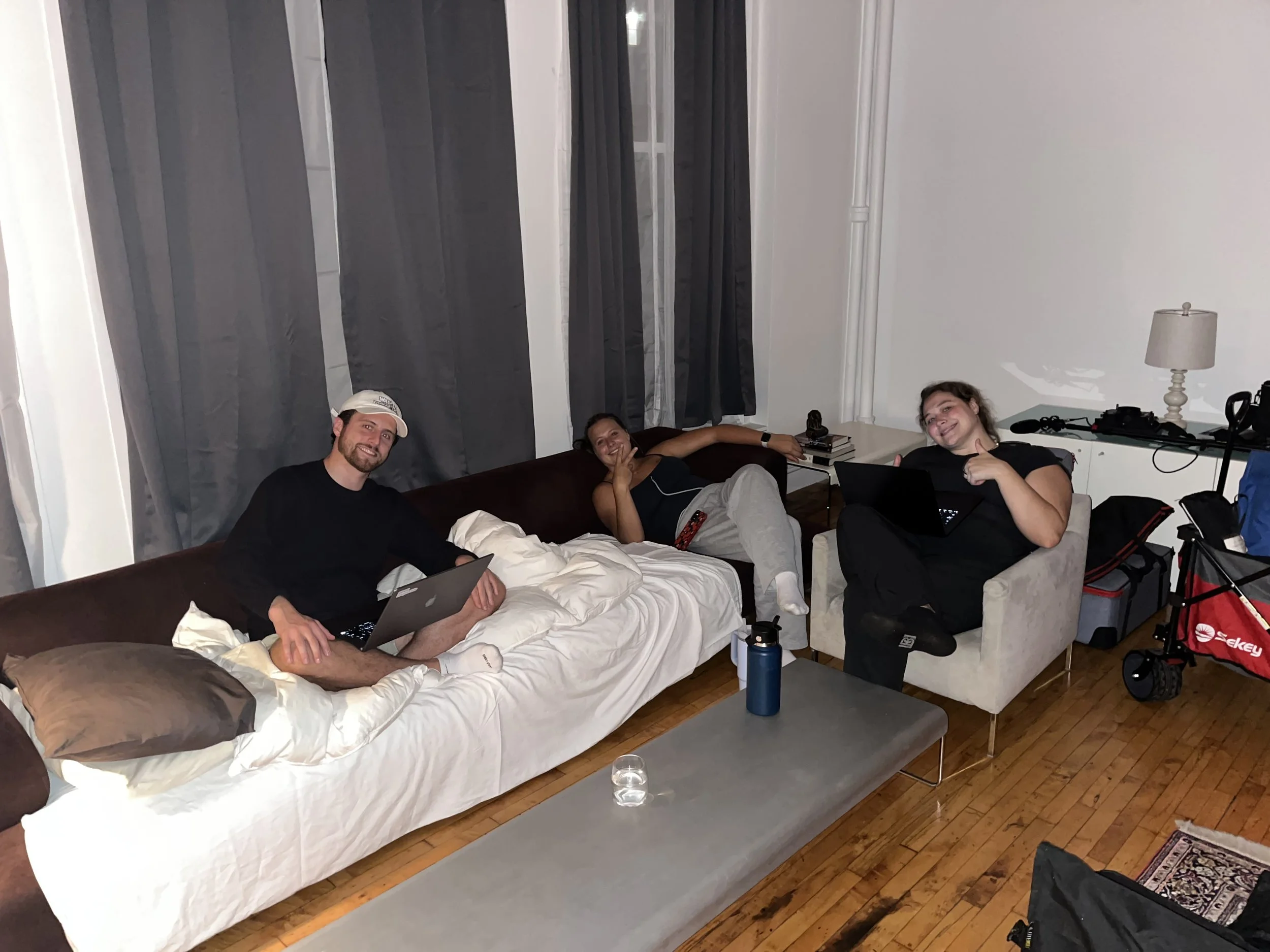 Three people are sitting in a room with gray curtains and hardwood floors, smiling and posing for the camera. One person is on a bed holding a laptop, another person is relaxing on the bed, and a third person is giving a thumbs-up, sitting on a chair