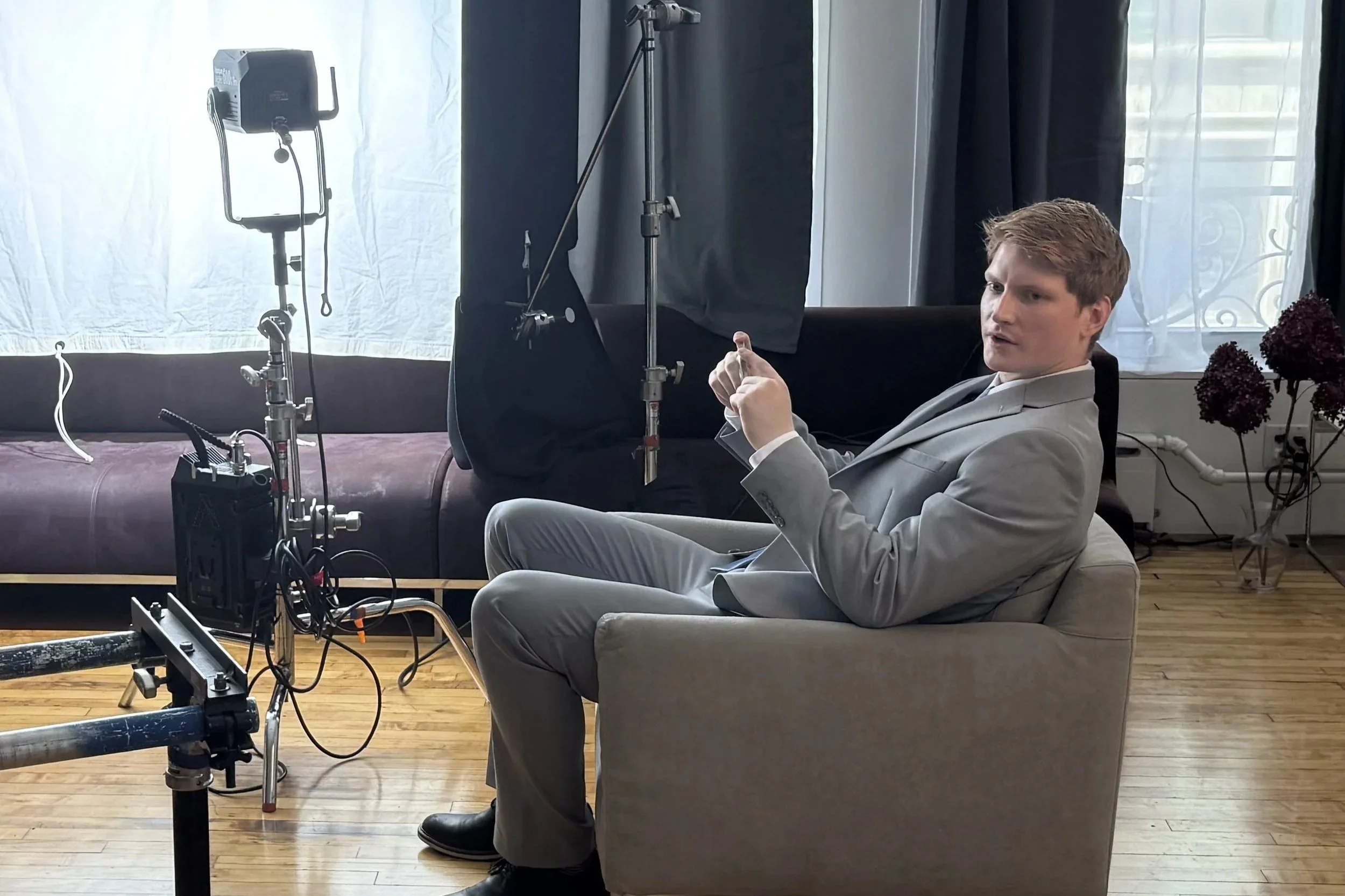 A man in a light gray suit sitting on a beige sofa in front of film equipment, with studio lighting and a white backdrop behind him.