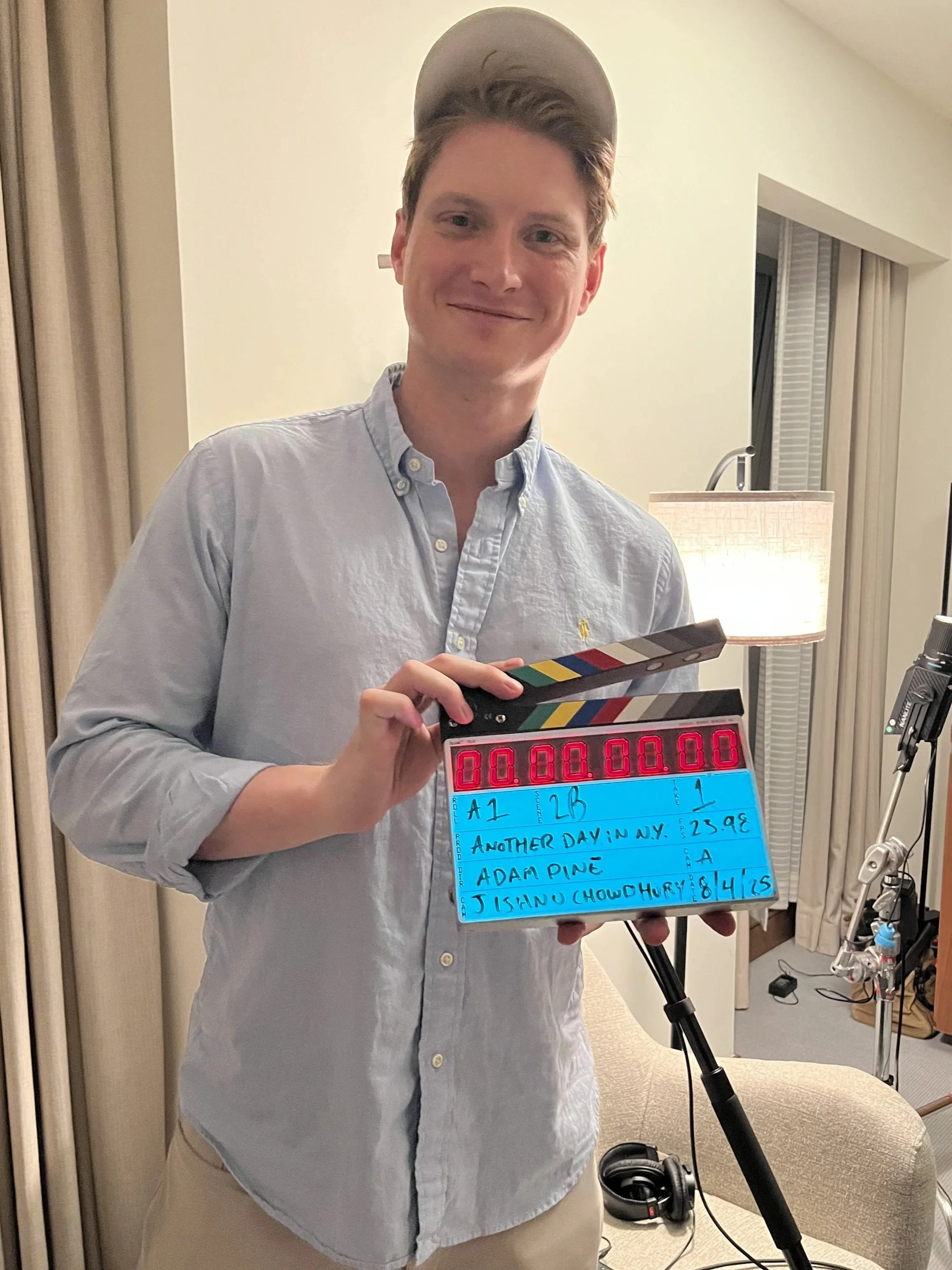 Adam Pine wearing a light blue button-up shirt and a gray hat smiling and holding a slate board with handwritten notes, in a room with beige walls, a lamp, a window with blinds, and photography equipment.