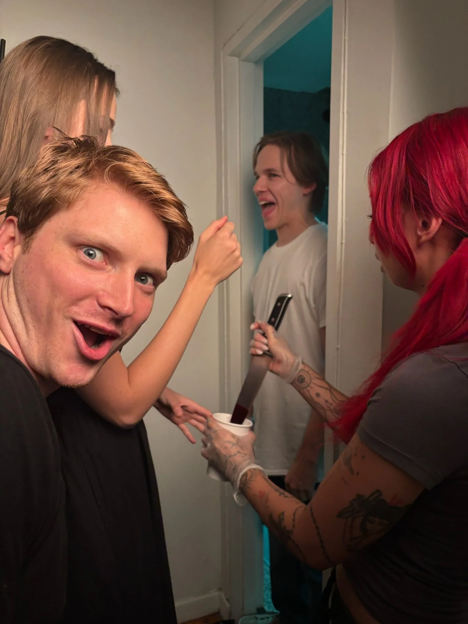 Group of people gathered in a room, with one person in the foreground looking excited and others engaging in a humorous or playful moment, including one person holding a large knife and another wearing colorful hair.