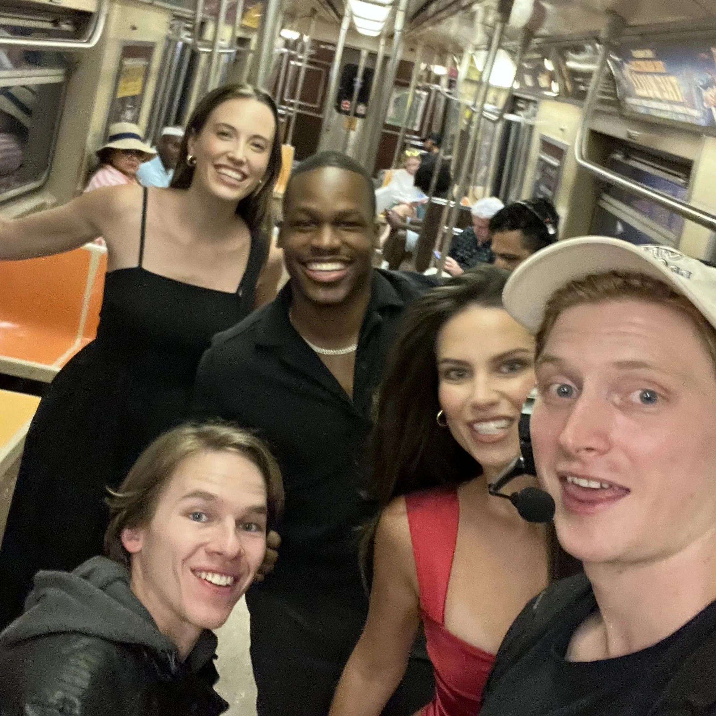 Group of excited people taking a selfie inside a train with beige walls, metal handrails, and windows showing a blurred scene outside.