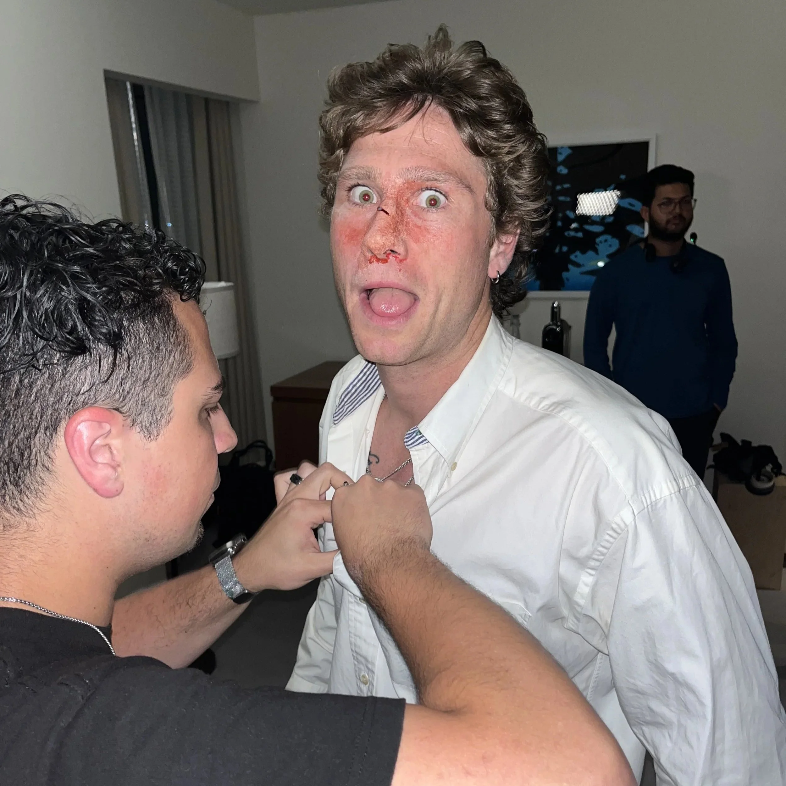 A man with wide eyes and an open mouth, appearing surprised or excited, is standing close to another person. The man has curly hair and a small earring, and he's wearing a light-colored shirt. There is a person with dark curly hair and a short haircu
