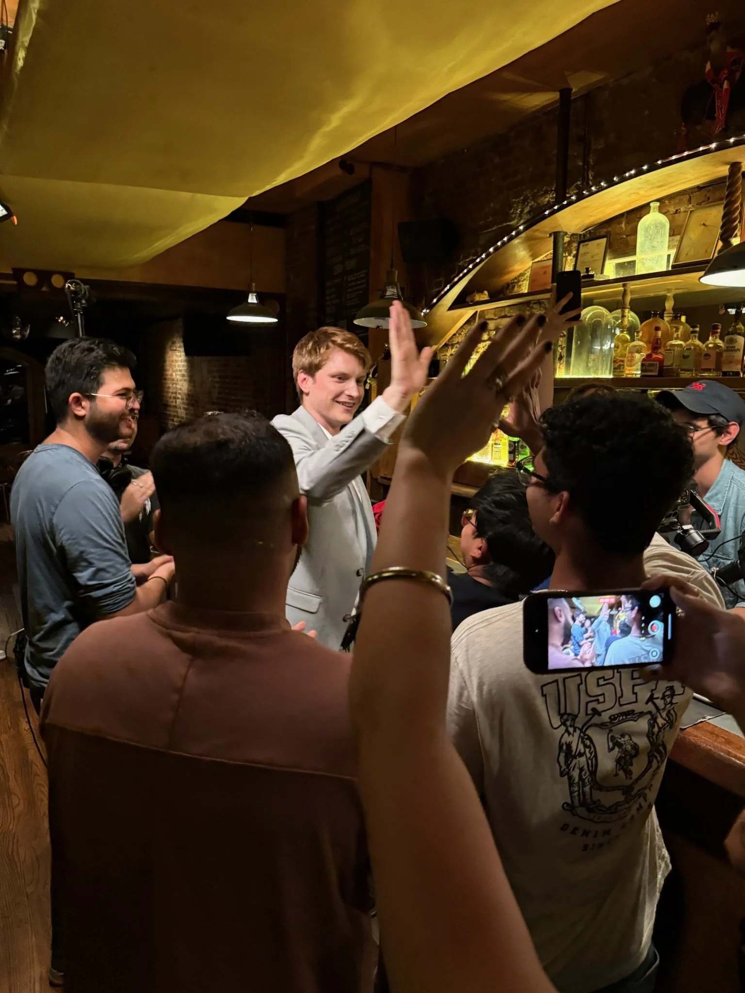 A group of people in a bar, some are holding drinks, one person in a gray blazer is raising their hand, and another person is taking a photo with a phone. The bar has bottles of alcohol on shelves illuminated with yellow lighting.