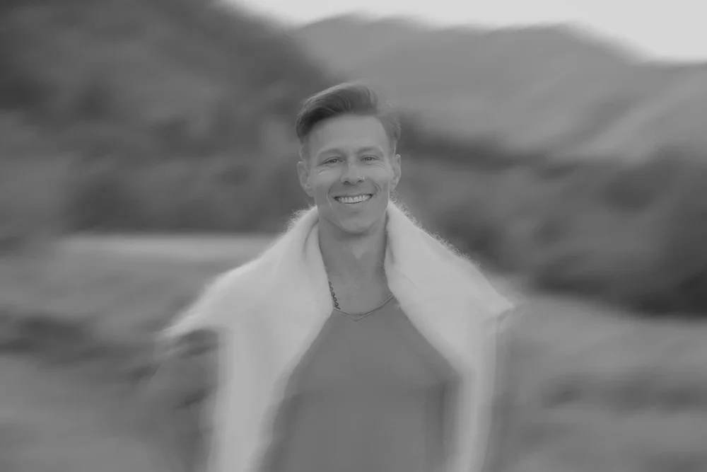 Black and white photo of a smiling woman outdoors with a scenic background of hills or mountains.